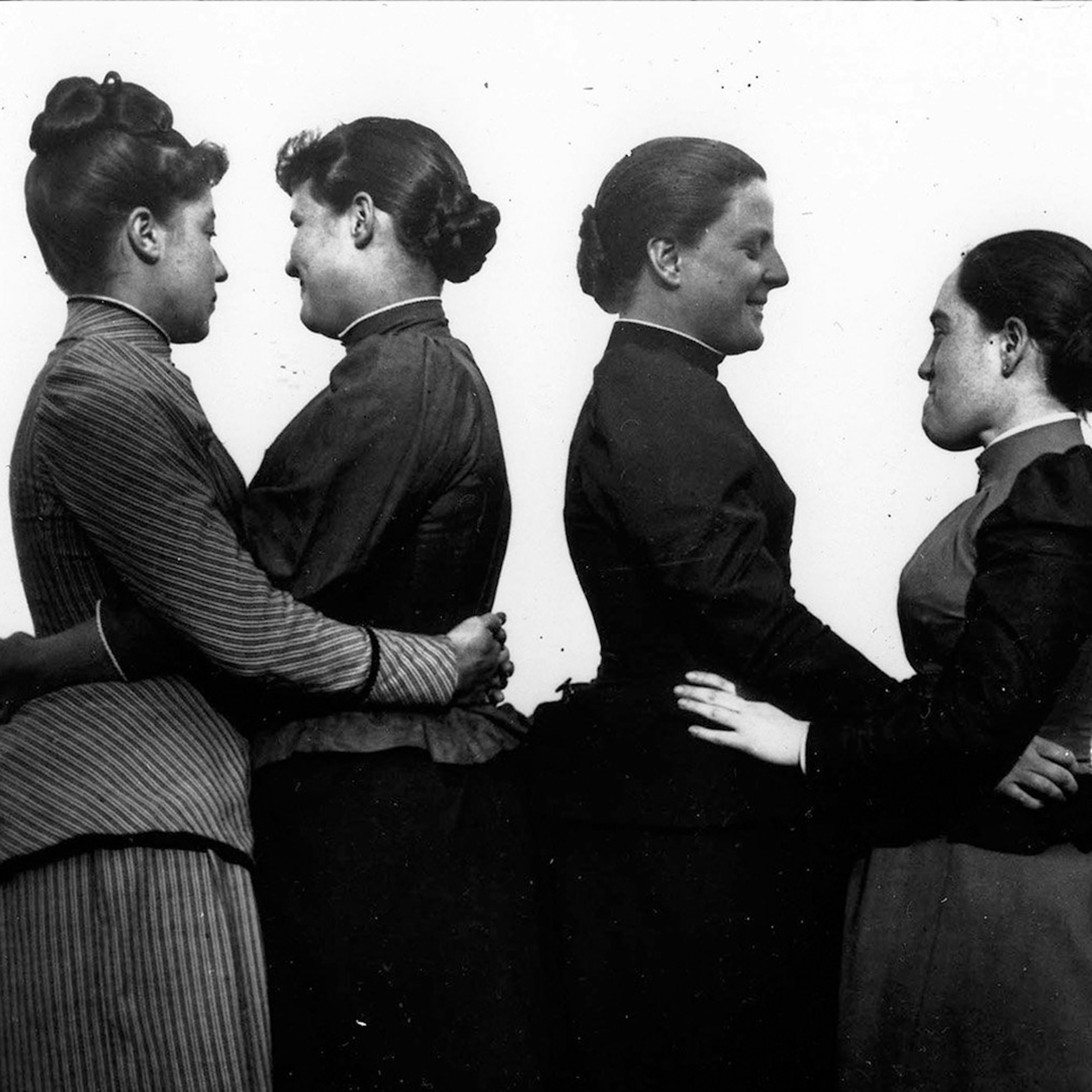 A black and white photo of four Victorian era women, they are paired off with each other and are hugging around the waist in a profile position compared to the camera.