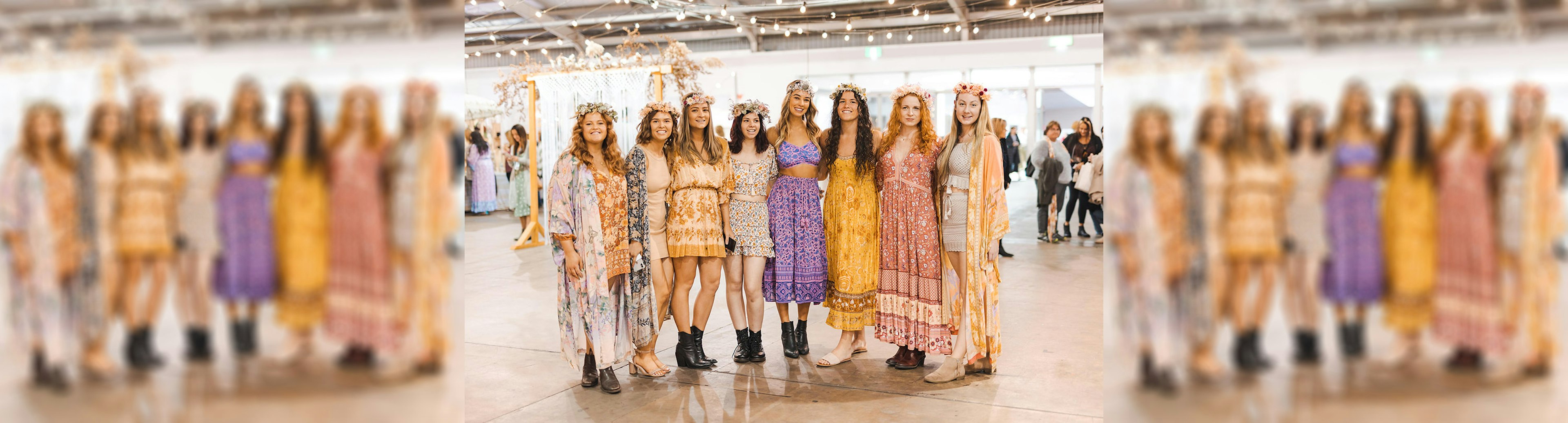 A group of smiling women with arms linked looking at the camera, they are all wearing flower crowns and are wearing dresses and skirts with flower patterns on them