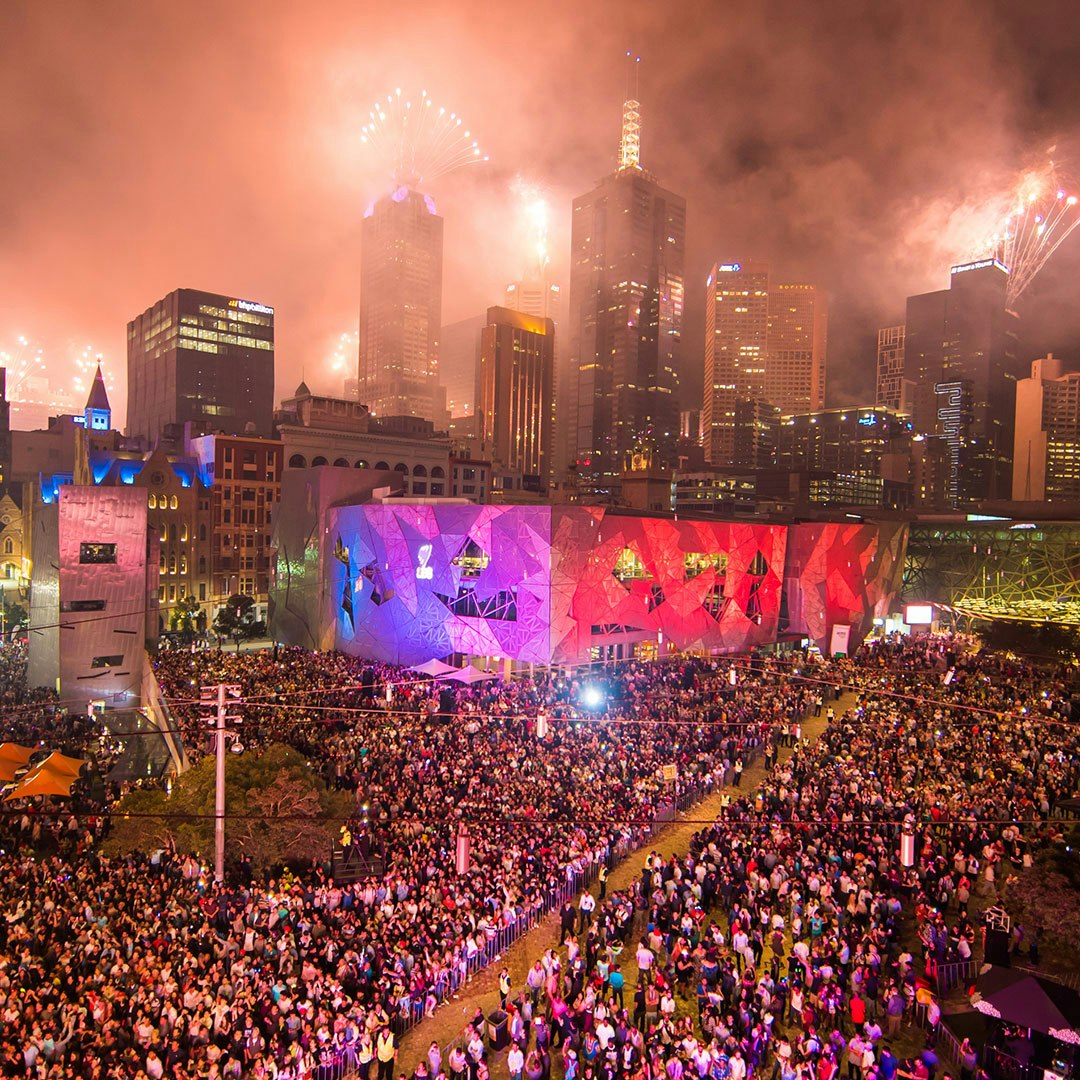 A photo of Fed Square on New Year's Eve as fireworks go off from rooftops in the background. The square is full of people