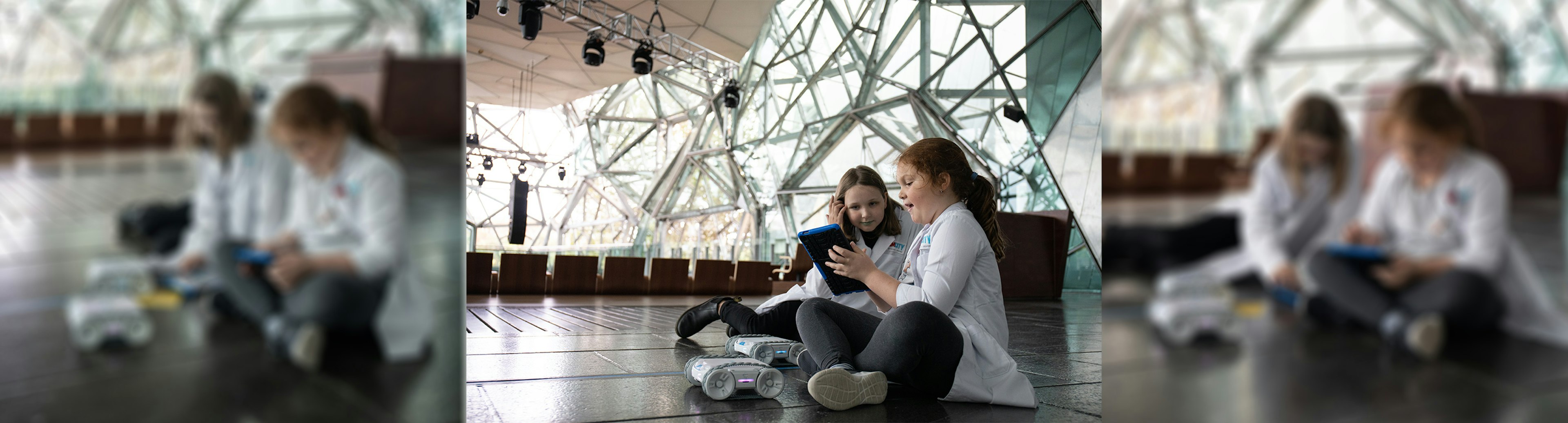 Two girls wearing white lab coats are sitting on the floor in The Edge at Fed Square, once of them is holding an iPad and they both looking at the screen. There are two remote control cars on the floor in front of them as well