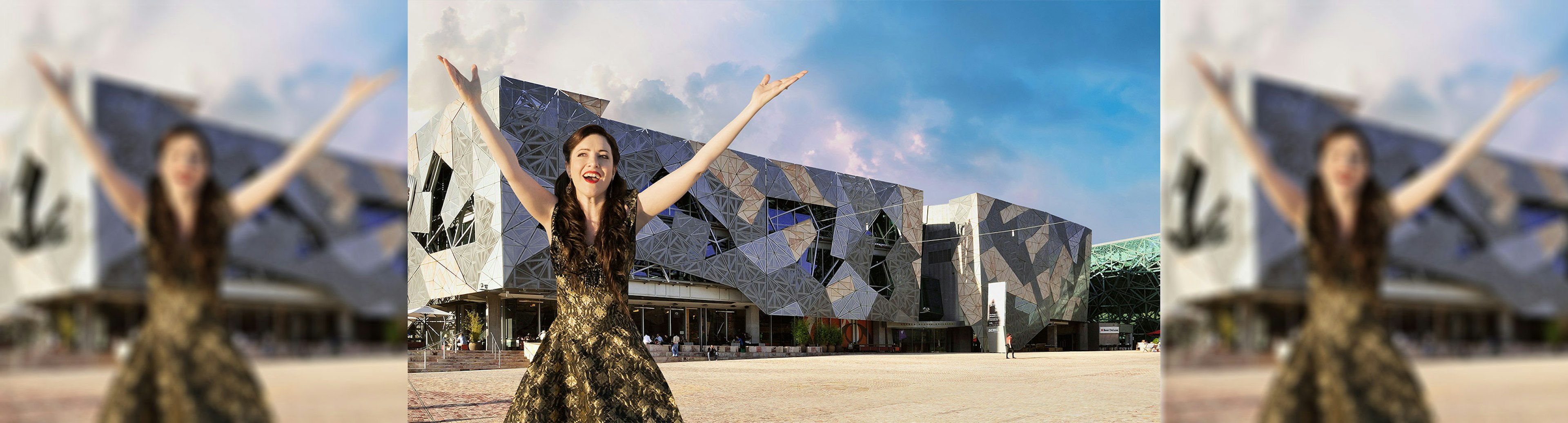 A woman in a black and gold dress with her arms open wide and up in the air, standing in front of a building in Fed Square