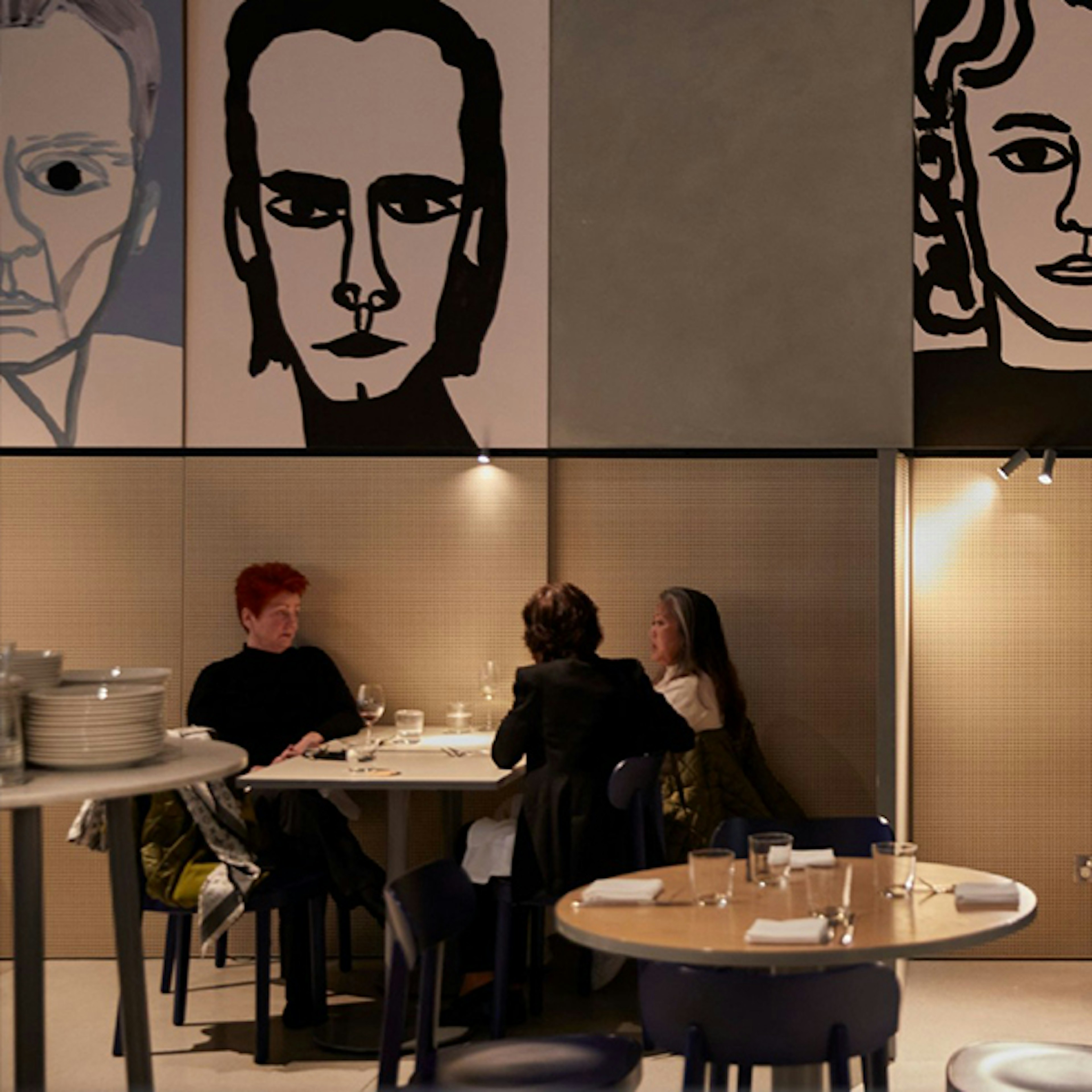 Three people sitting at a table up against a wall at HERO, there is an obscure portait of Nick Cave above them