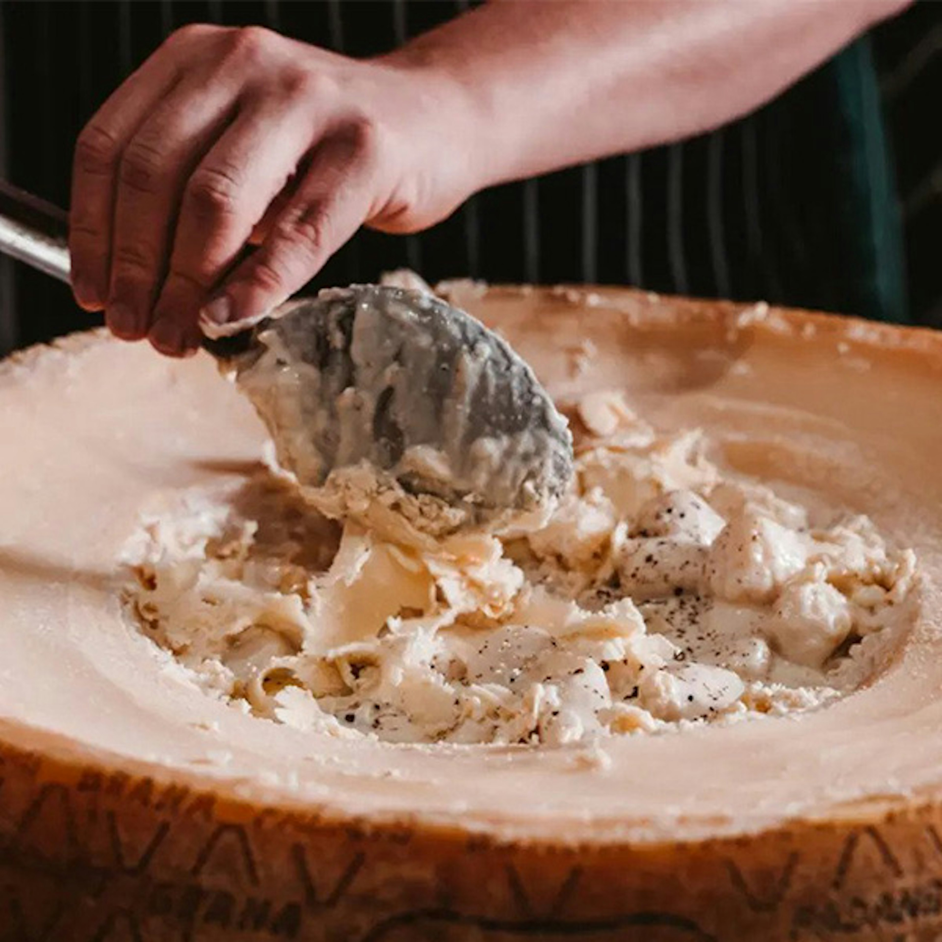 A close up of a big wheel of cheese with a person scooping out the inside with a spoon