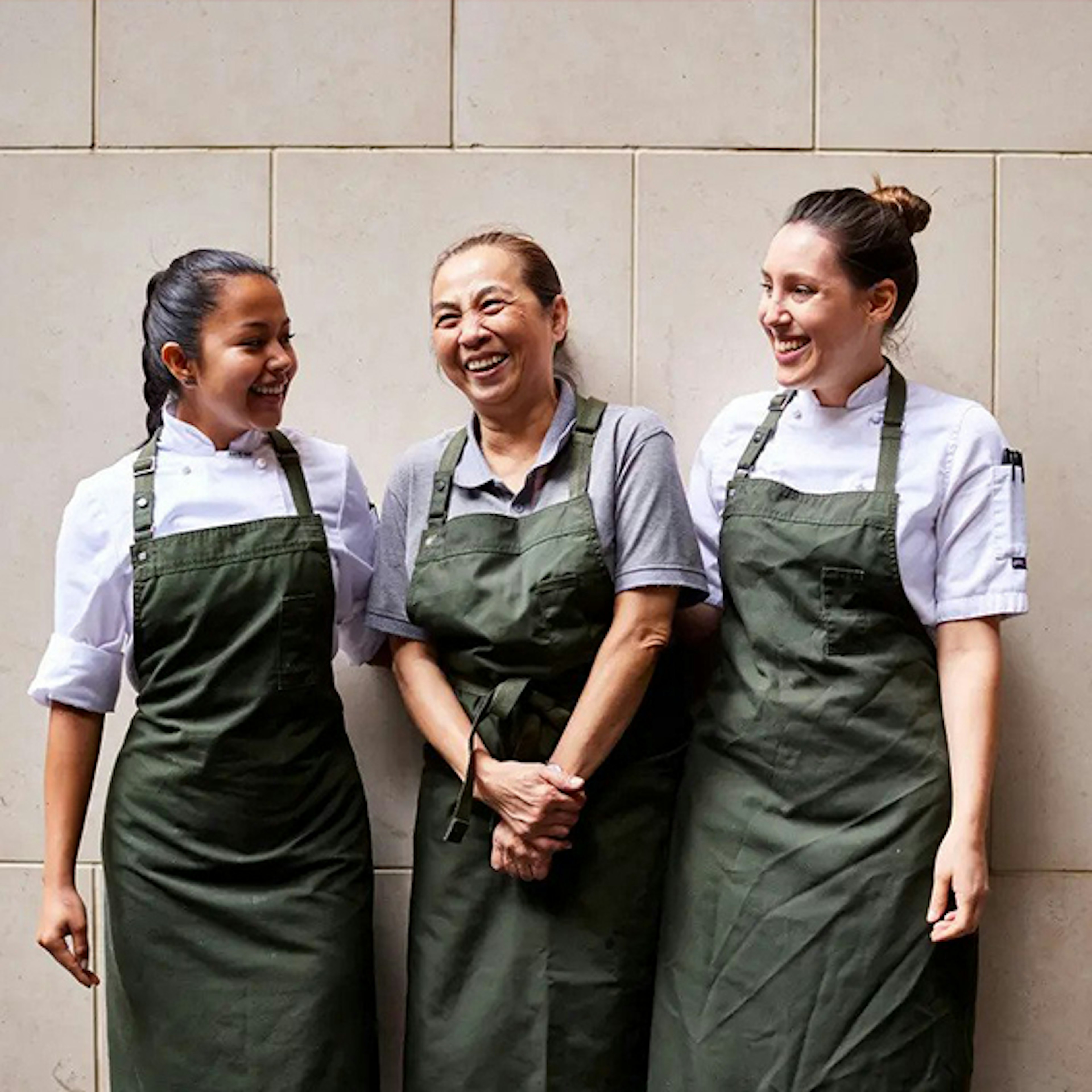 Three female chefs standing against a wall and smiling at one another, they are all wearing a dark green apron with chef whites underneath