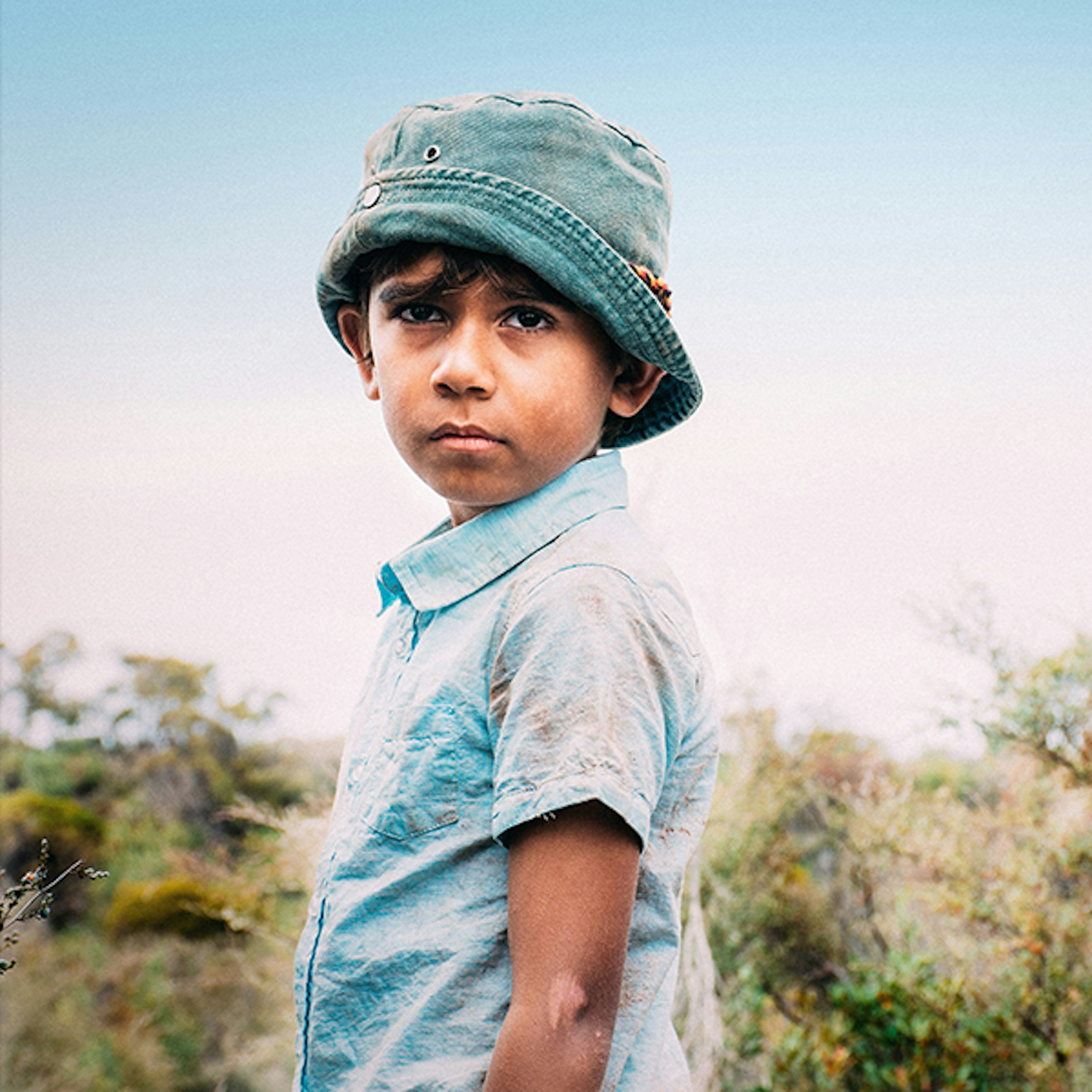 A young boy wearing a blue bucket hat and light blue collared shirt is standing in the scrub profile, looking at the camera