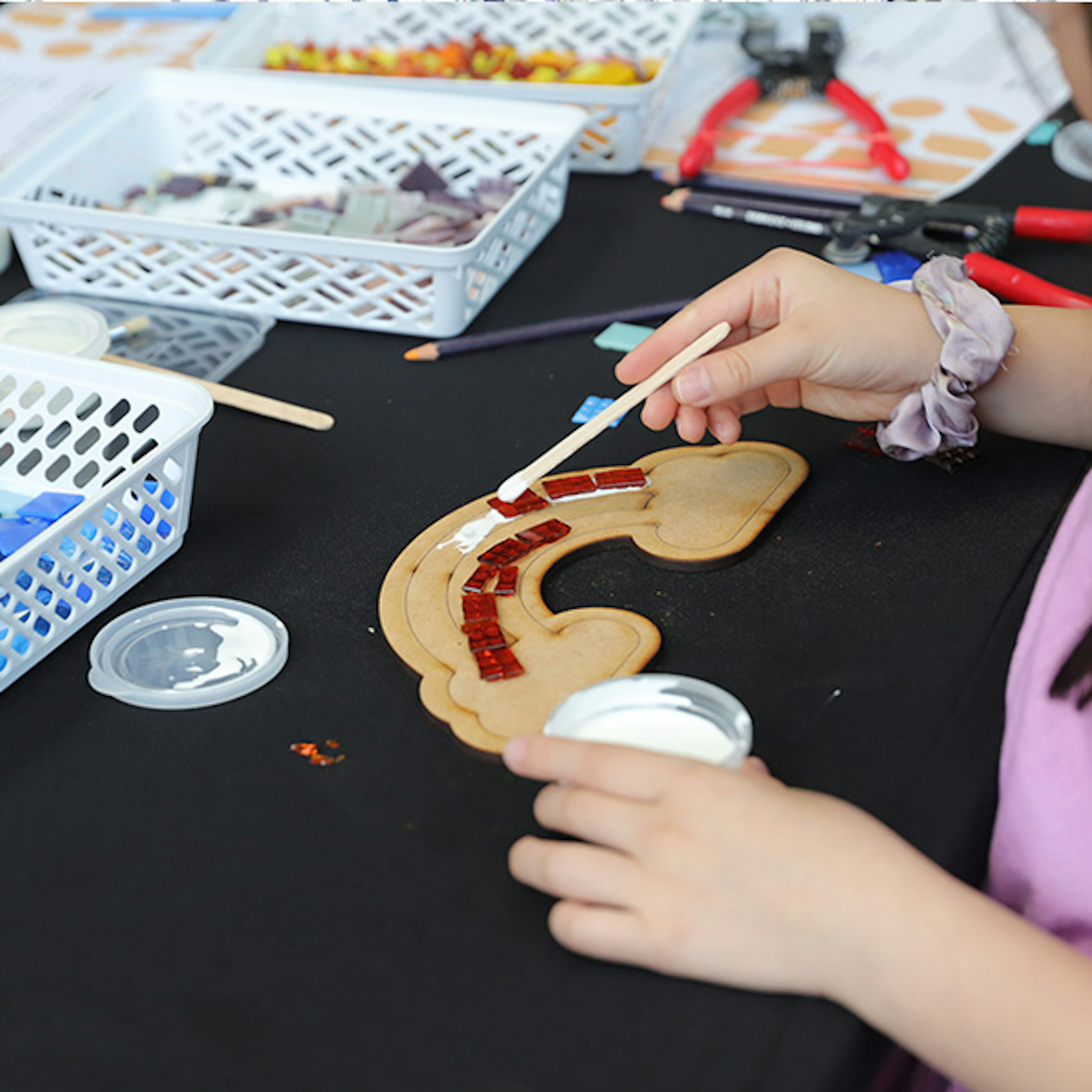 A child that is just out of the shot is creating a mosaic rainbow with red tiles and PVC glue