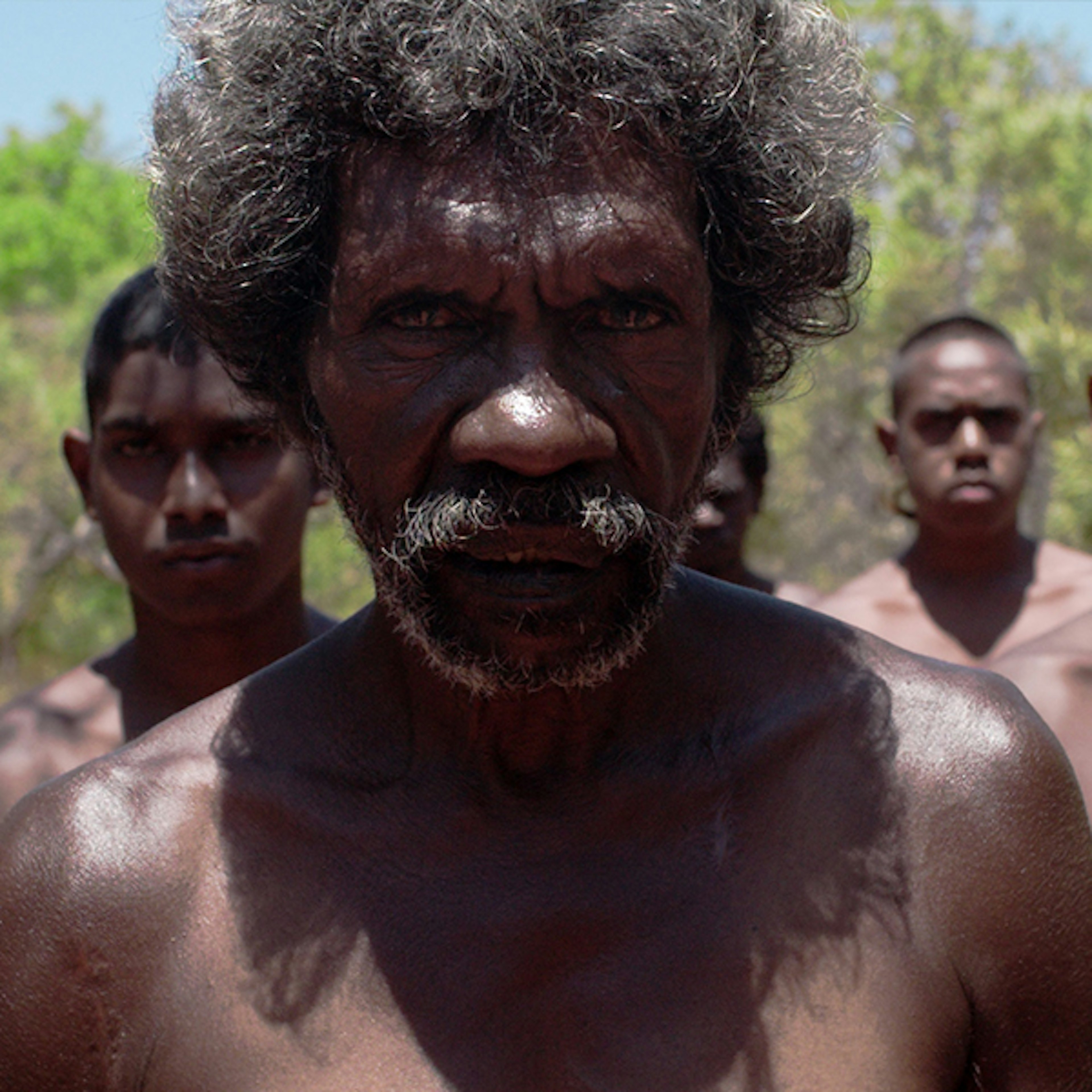 An Aboriginal Australian man with greying hair and moustache is wearing no shirt and staring into the camera with no expression