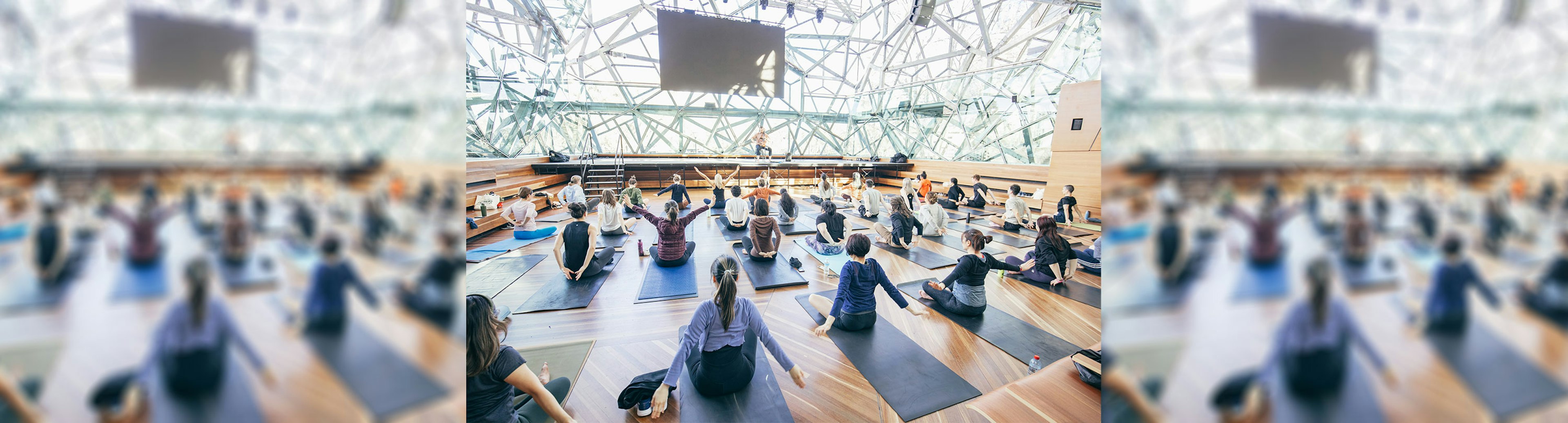 A room full of people sitting on mats doing yoga while an instructor is up on a stage