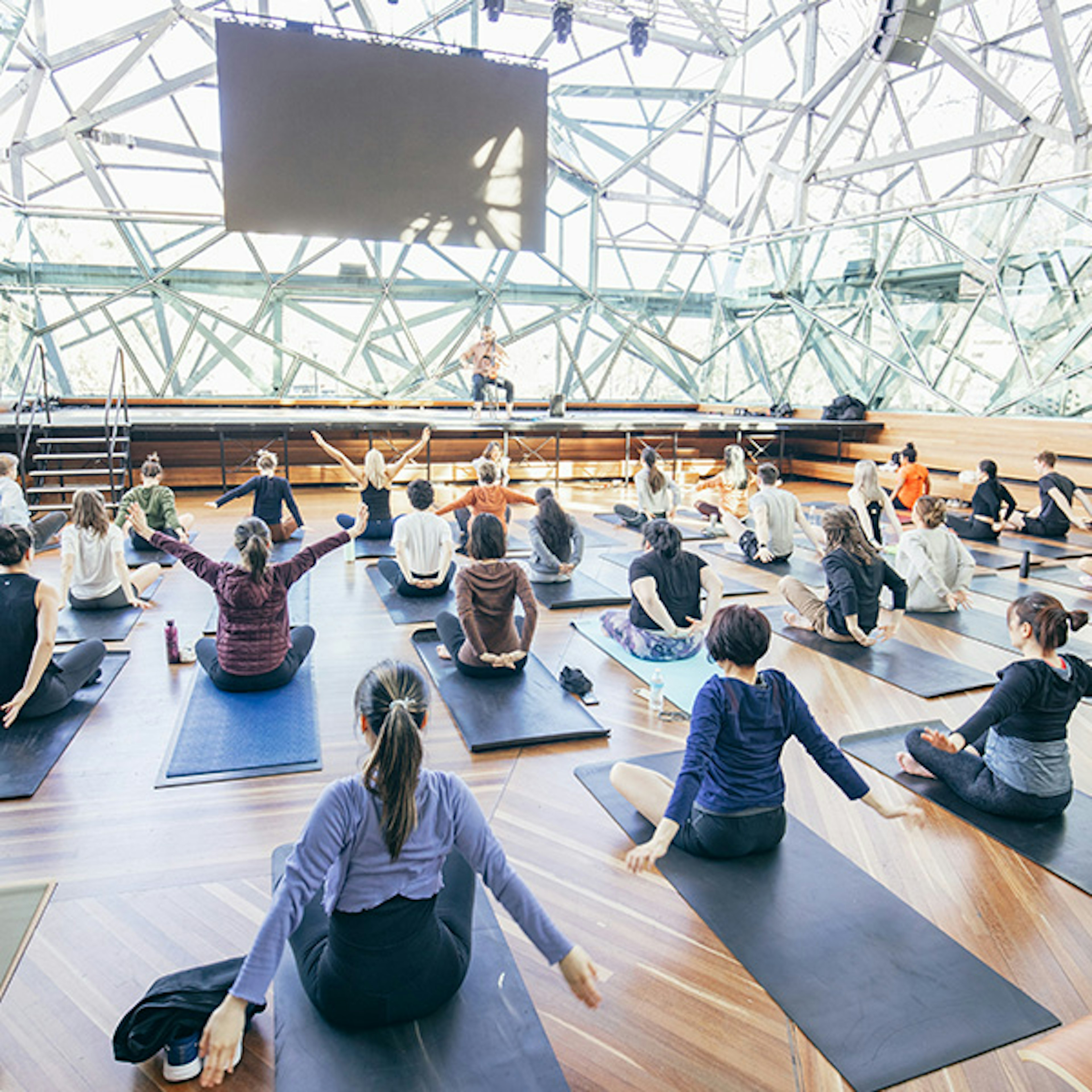 A room full of people sitting on mats doing yoga while an instructor is up on a stage