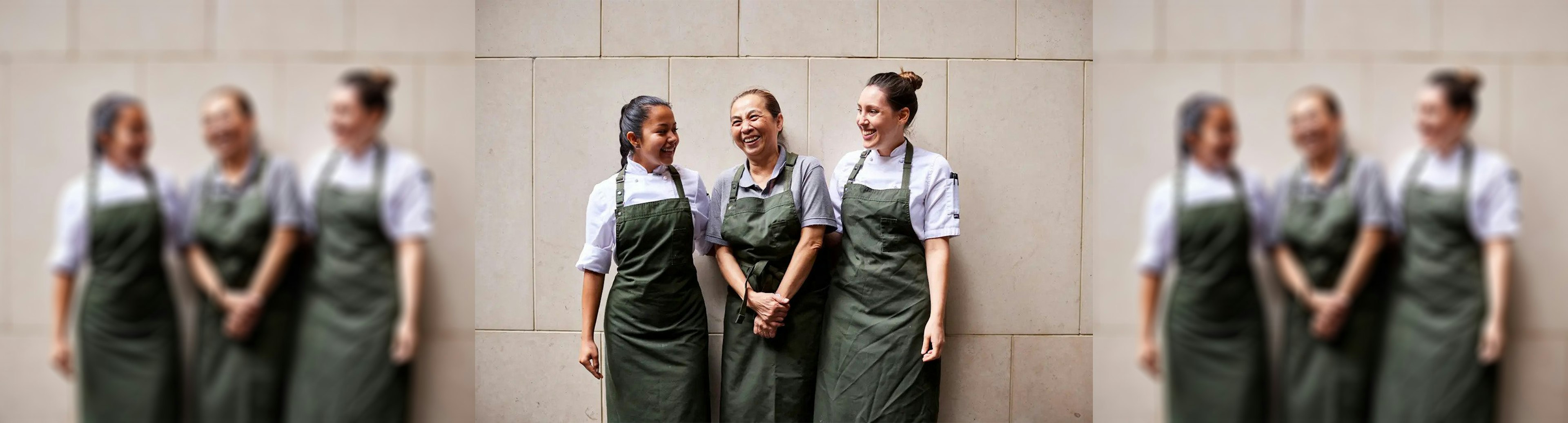 Three female chefs standing against a wall and smiling at one another, they are all wearing a dark green apron with chef whites underneath