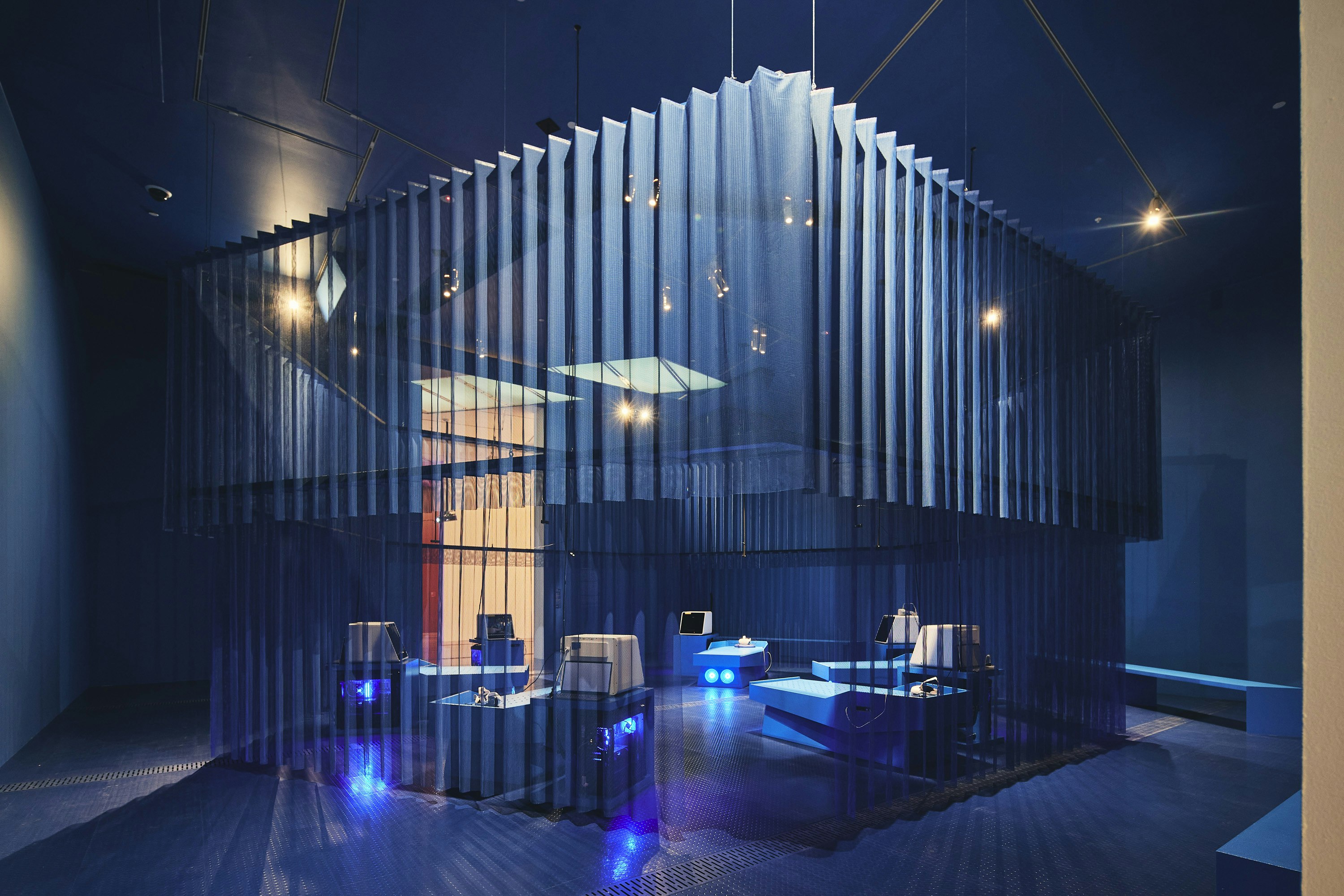 A see-through curtain encloses a square space within a large gallery room. Metal beds are within the room, and there is blue light coming from machines next to the beds.