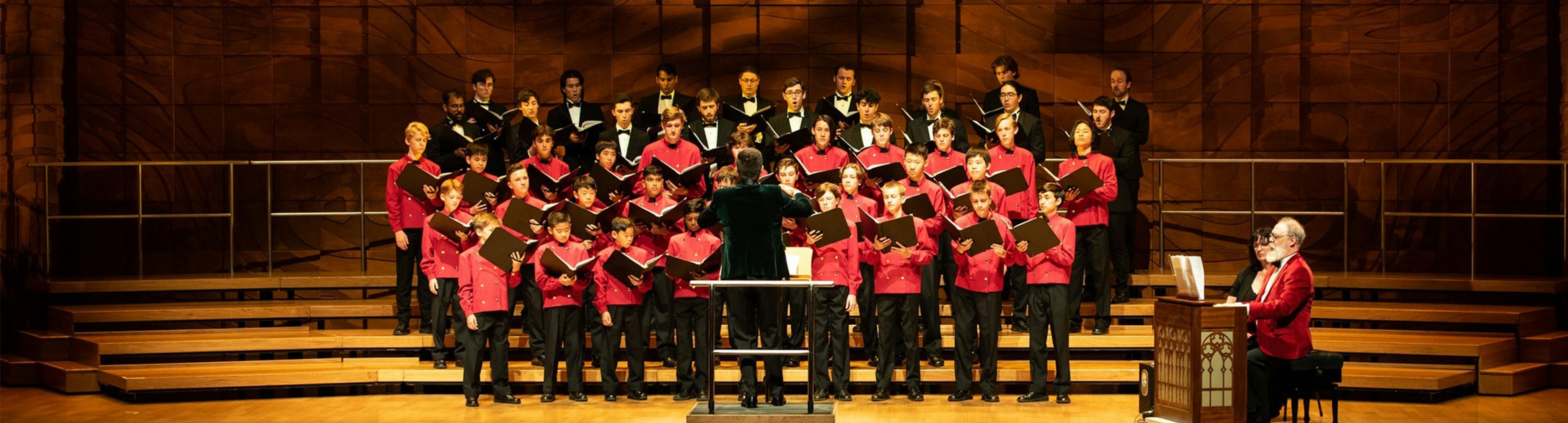 The Australian Boys Choir on stage performing, the first three rows are wearing red jackets and the top two rows are wearing tuxedos, all of them are holding open folders looking at the words they are singing. A conductor has their back to the camera standing in front of the choirs