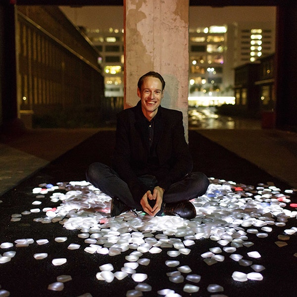 Dutch artists Daan Roosegaarde is sitting on the ground in front of a concrete pillar wearing black clothing and smiling at the camera