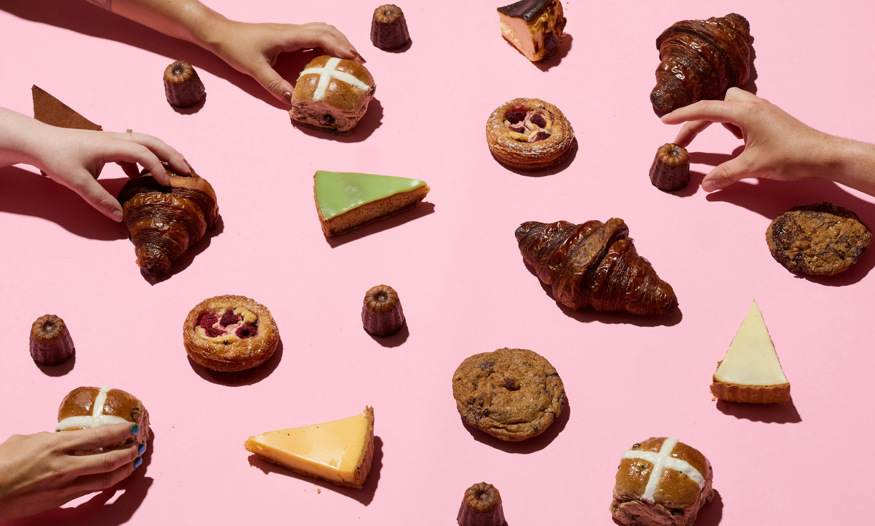 Lots of different pastries sitting on a light pink background with three hands coming in from the left grabbing three different items