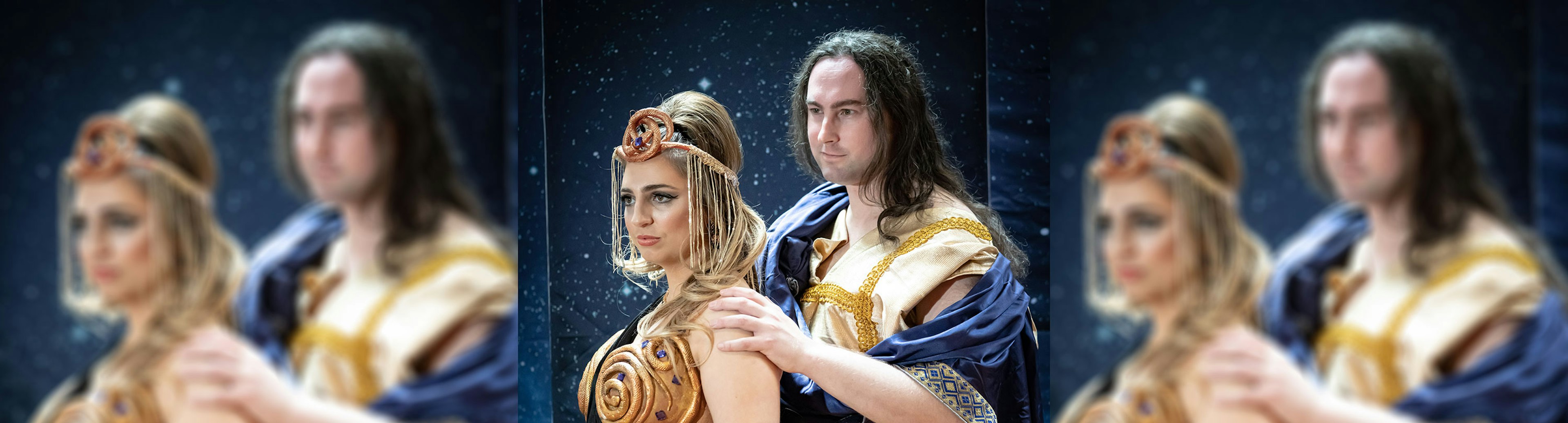 Two people are posing for an opera performance wearing ancient Roman style clothing, a man with long dark hair is standing behind a woman with long blonde hair and his arm is resting on her shoulder.