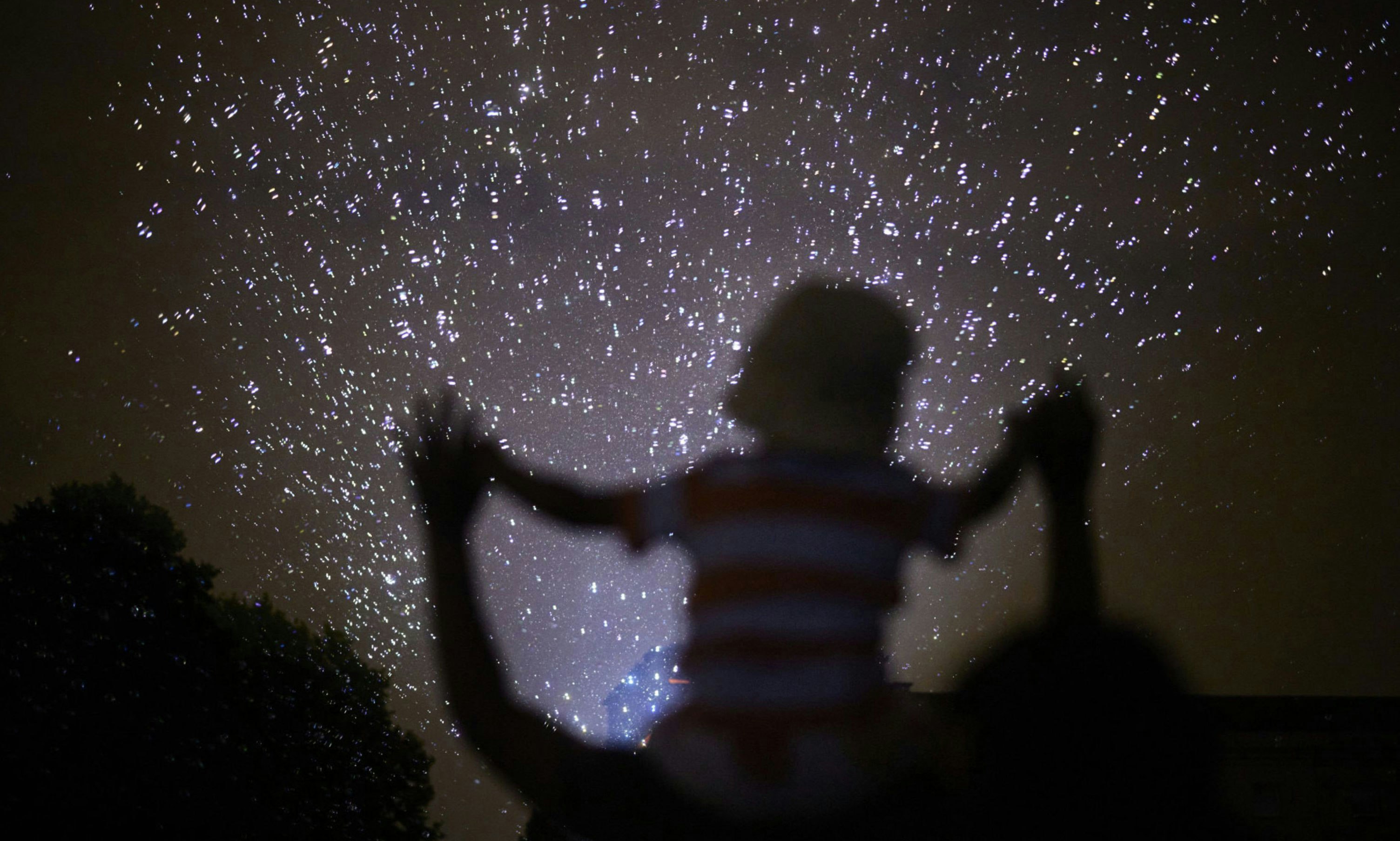 An outline of a child on an adults shoulders as they are looking up into the night sky at lots of small lights
