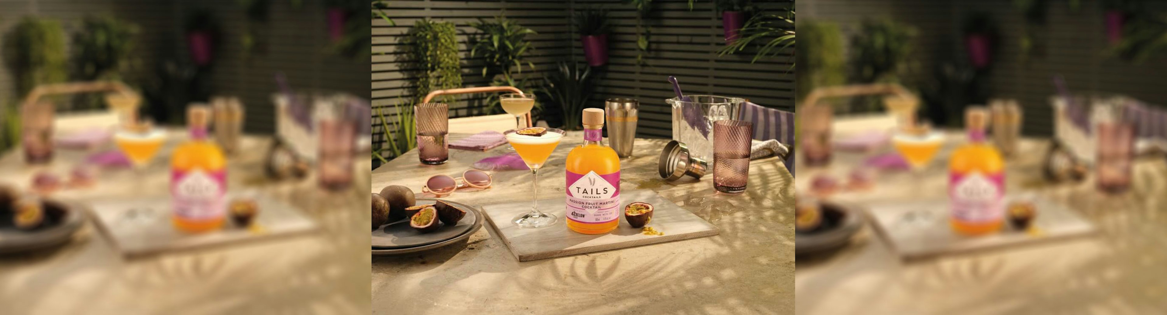 A bottle of TAILS' pre-mixed passionfruit martini sitting on a table surrounded bt a martini glass, shaker, ice bucket, sunglasses and passionfruit