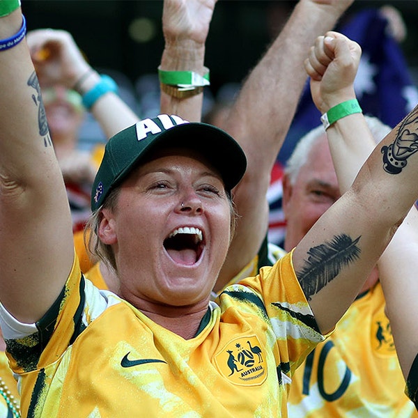 An Australian soccer fanning wearing the green and gold jersey smiling with their arms up in the air in celebration