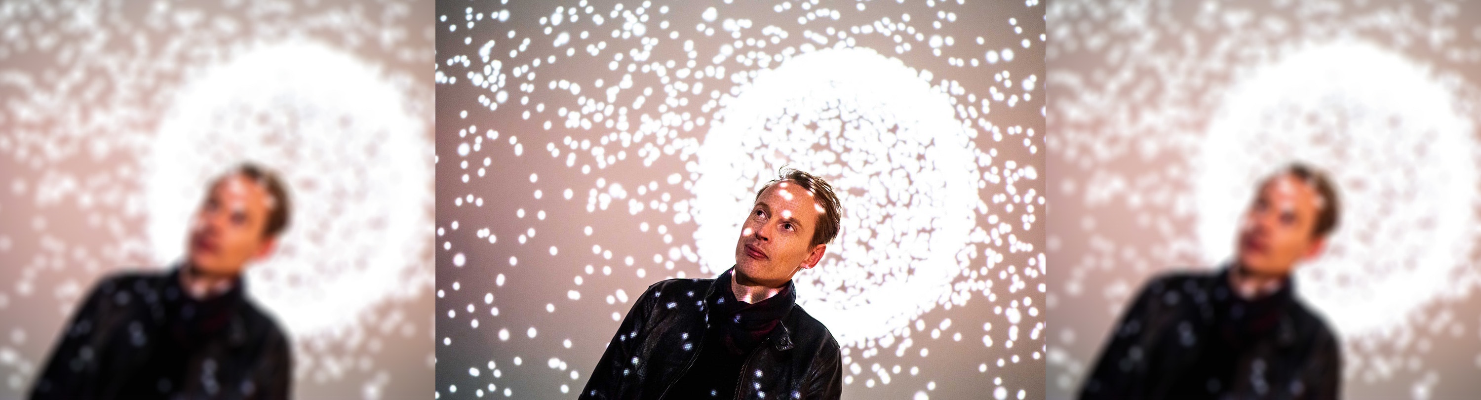 Artist Daan Roosegaarde is standing in front of a projection of a spark of sparkling lights and is wearing a dark leather jacket