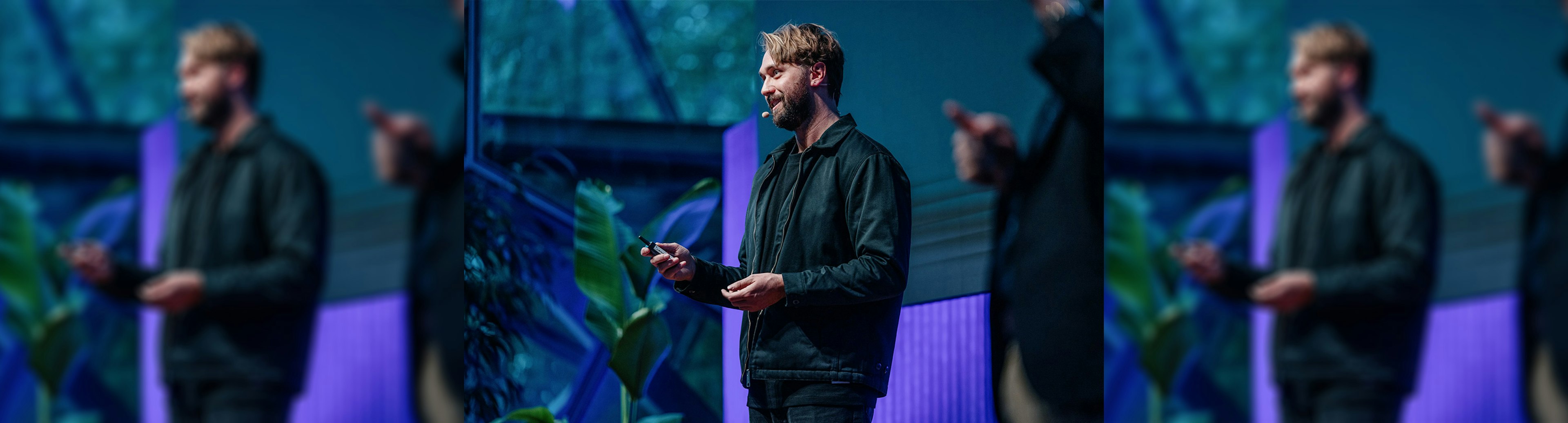 A man wearing a black jacket is talking to a crowd while on stage, and is holding a clicker