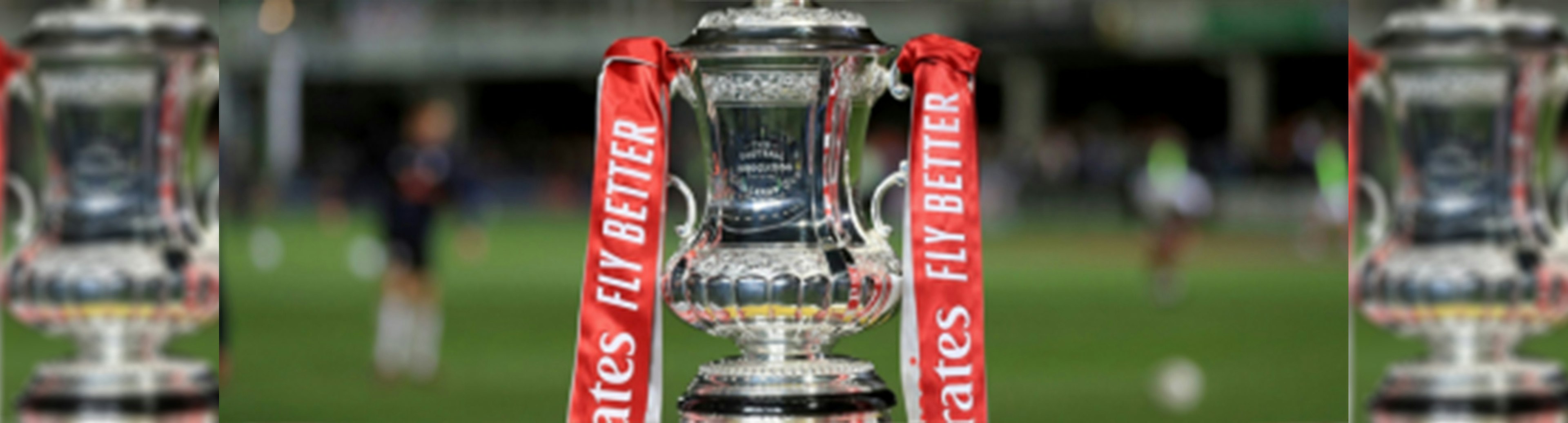 The FA Cup Trophy is front and centre with grass of a pitch and soccer players blurred in the background