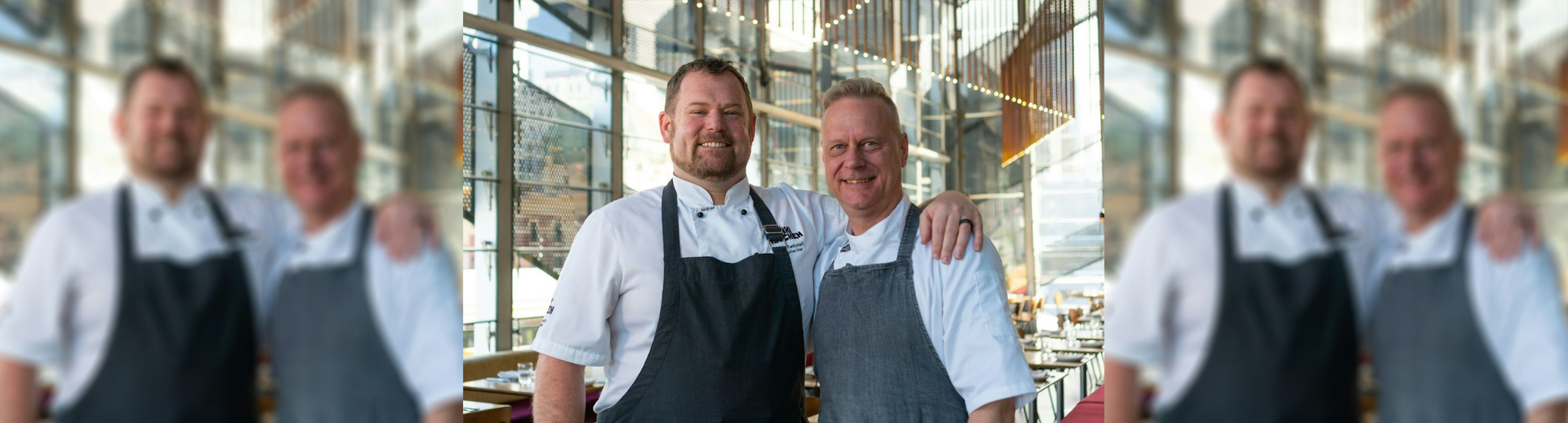 Two chefs are standing side by side wearing chef whites with aprons over the top. Both are smiling at the camera and the chef on the left has their arm around the shoulder of the chef on the right