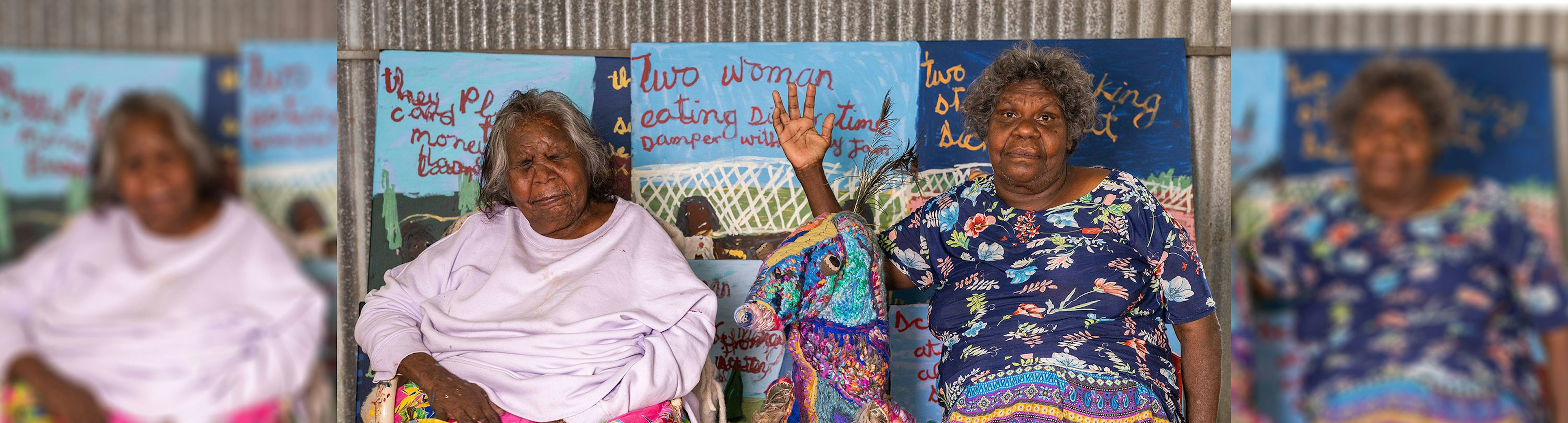 Artists Sally M Nangala Mulda and Marlene Rubuntja are sitting on seats next to each other facing the camera. Sally has her eyes closed and Marlene has her right arm up and is looking directly at the camera.