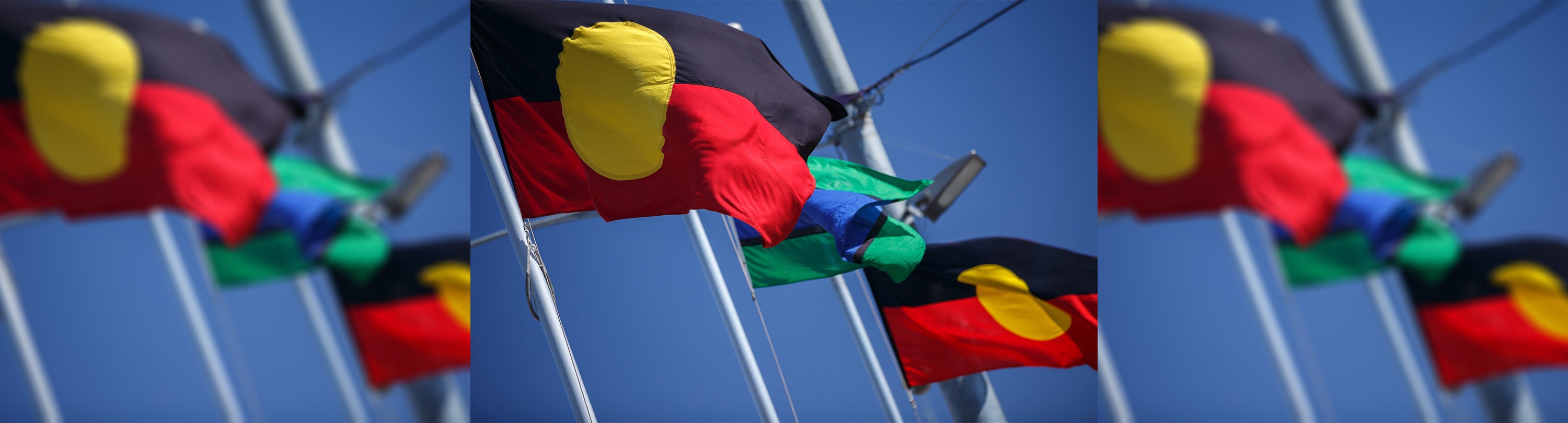 The Aboriginal and Torres Strait Island flags are waving in the wind on flag poles in front of a blue sky