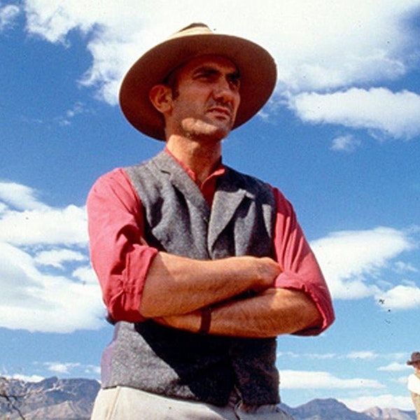A film still of singer and songwriter Paul Kelly in the film One Night the Moon. He is standing with is arms crossed wearing an Akubra hat a light read shirt with the sleeves rolled up and a grey cotton vest over the top