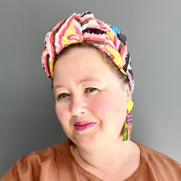 A woman with a colourful head band is facing the camera and smiling with her head slightly tilted to the left so that you can see her earring