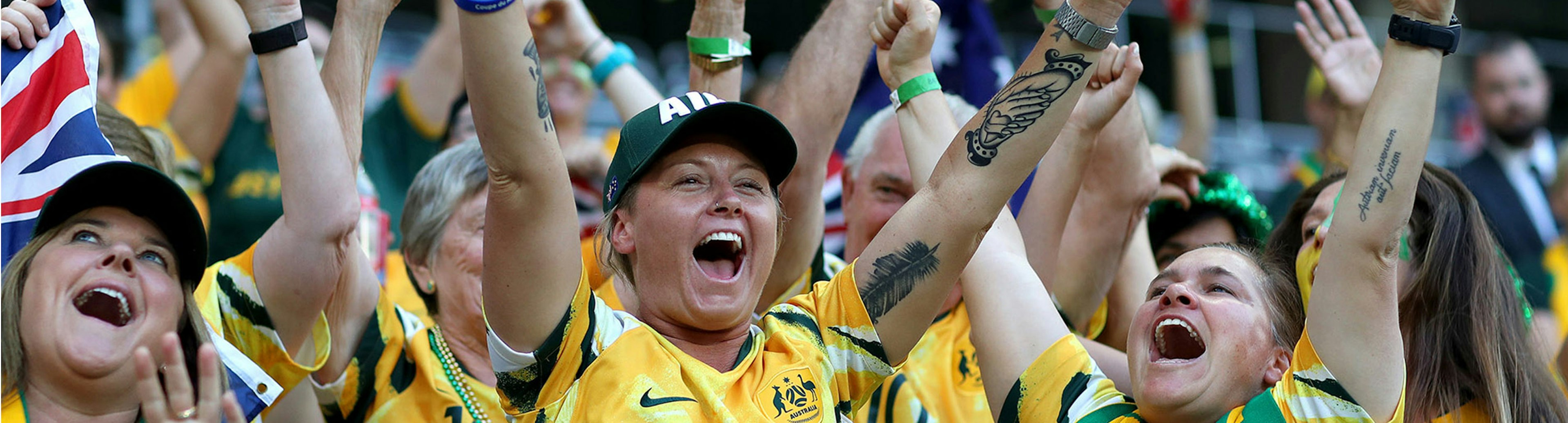 An Australian soccer fanning wearing the green and gold jersey smiling with their arms up in the air in celebration