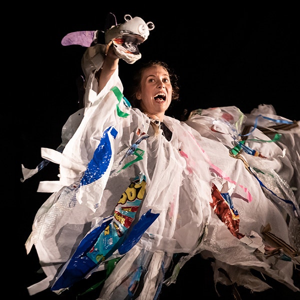 A woman performing with a puppet on her hand made from trash and wearing an outfit made from trash as well