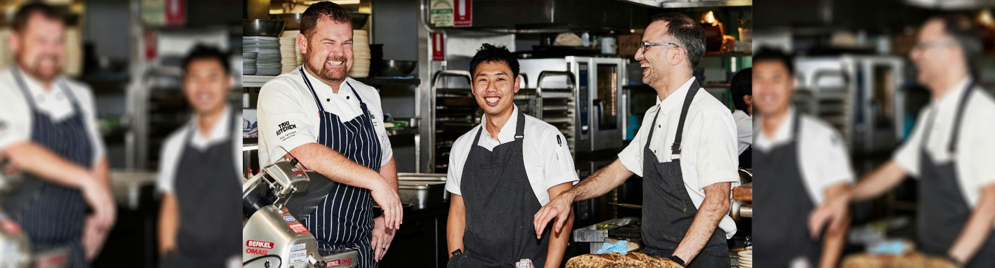 Three chefs in a kitchen wearing whites and blue aprons smiling at each other