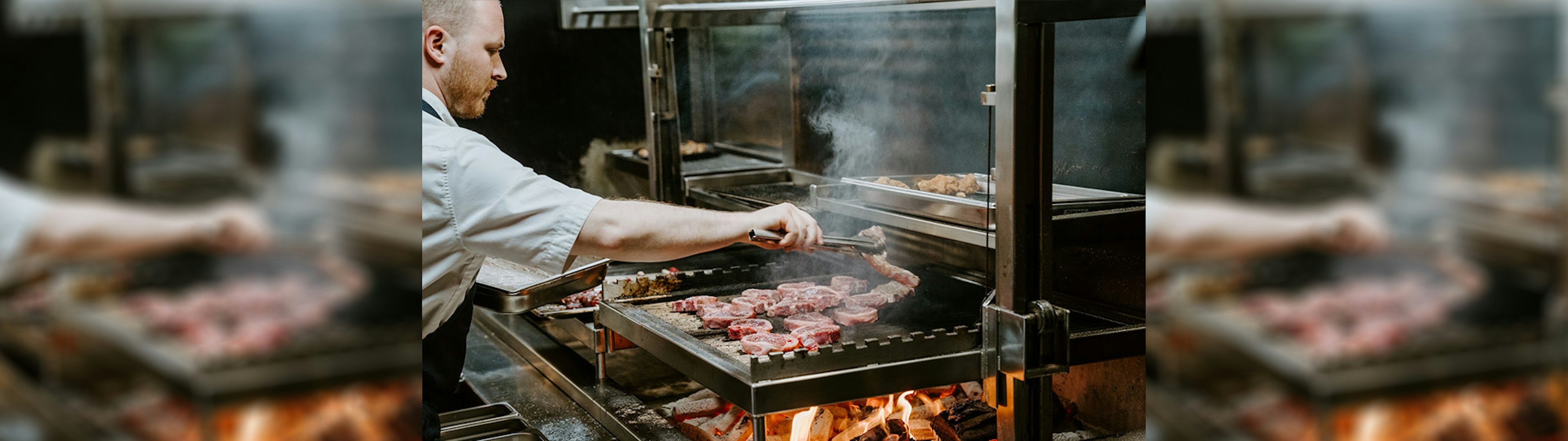 A chef leaning over a wood-fired grill putting lamb chops onto the grill with tongs