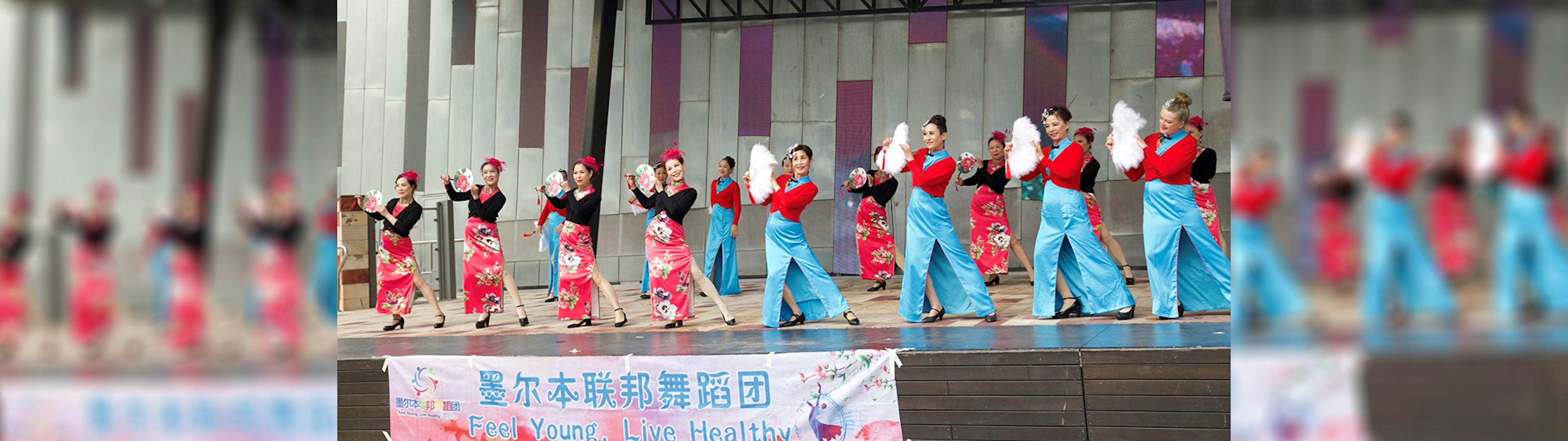 A troupe of gentle Chinese dancers wearing colourful qipoa are dancing on the Fed Square main stage