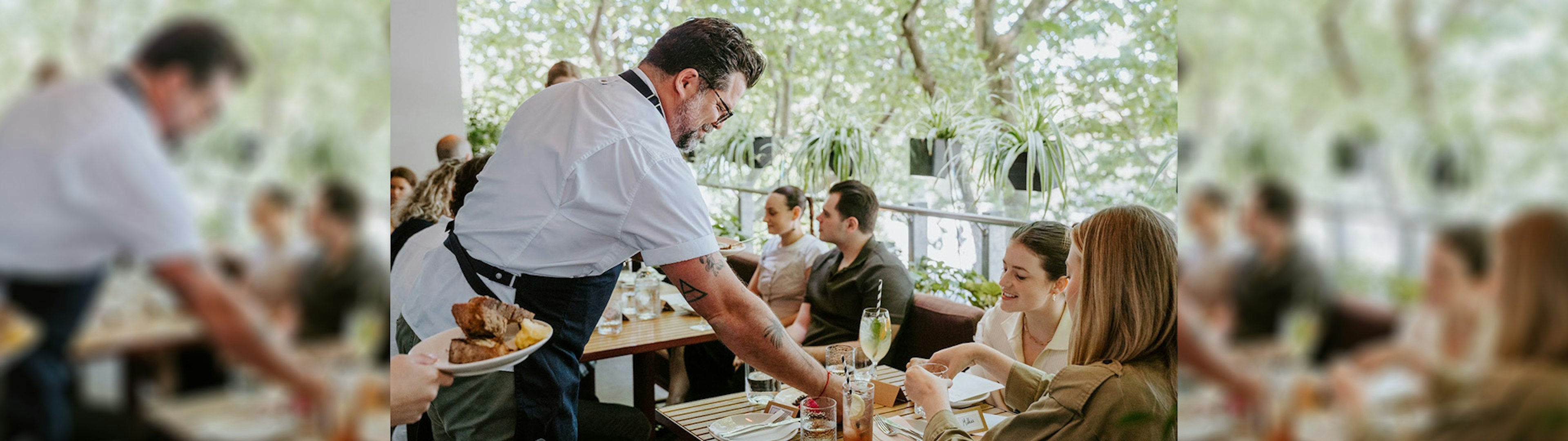 A chef is serving customers food and drinks on a table on the terrace at Victoria by Farmer's Daughters