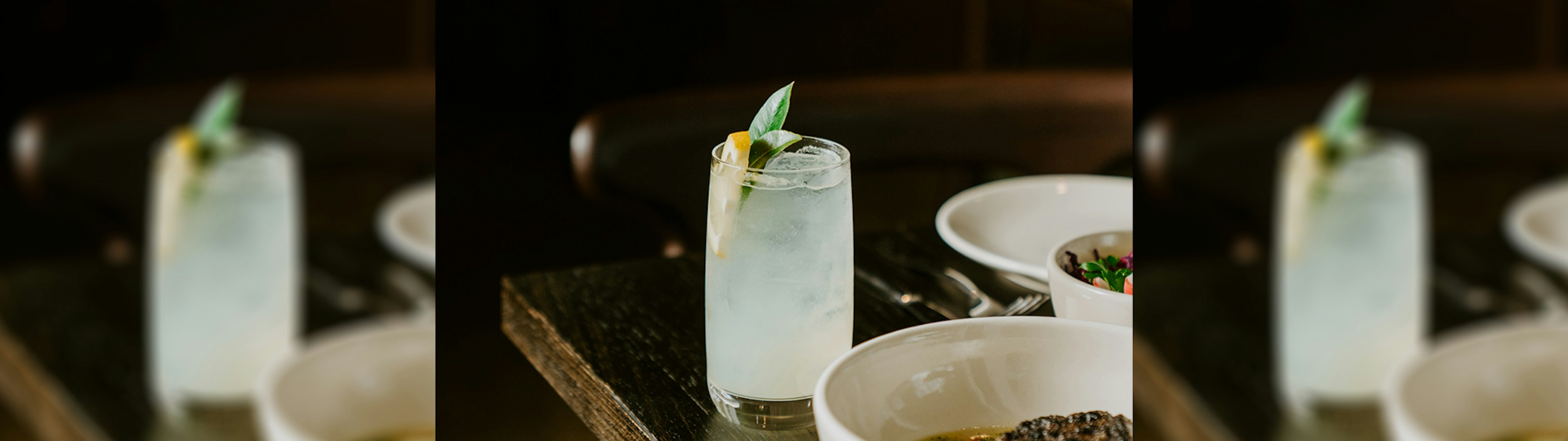 A cocktail on a table next to a plate, the cocktail is gin based but looks like a refreshing lemonade