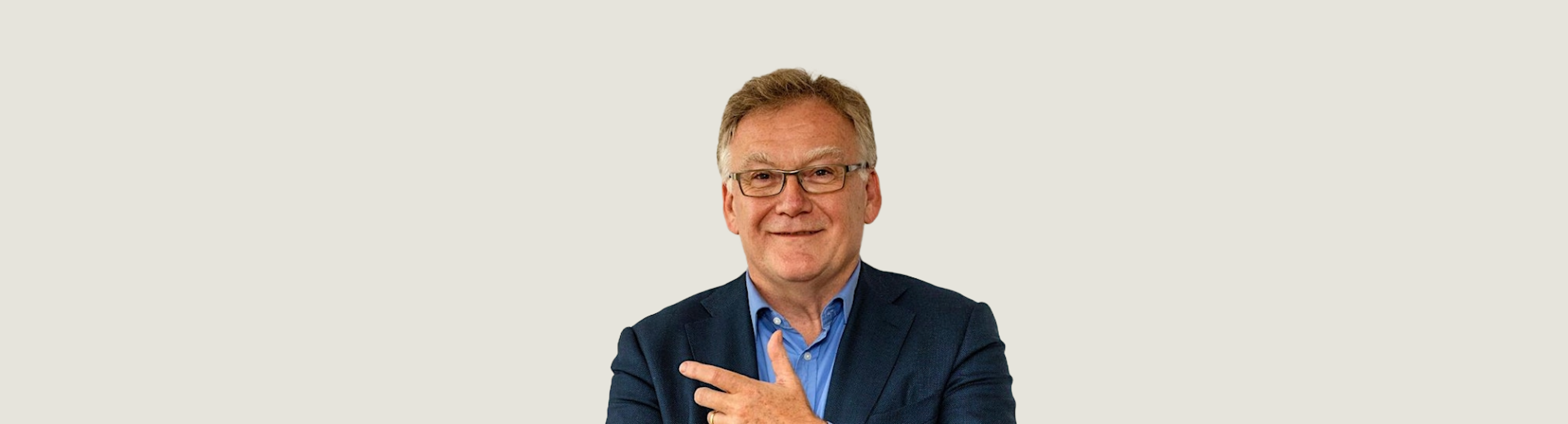 A middle aged man wearing glasses and a blue jacket is standing in front of a cream wall and smiling directly at the camera