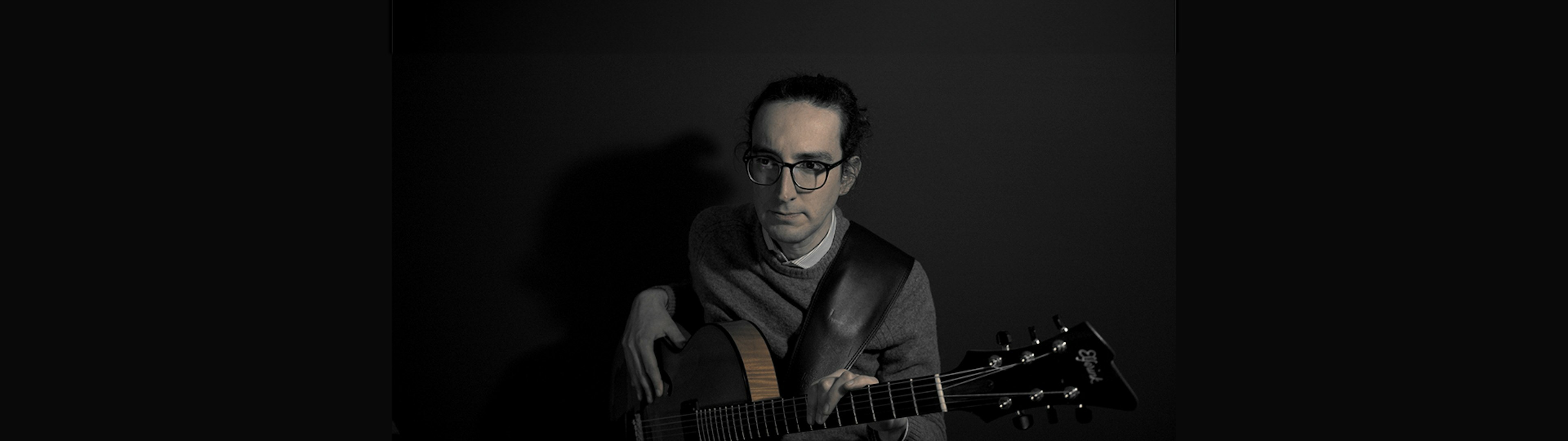 Jazz guitarist Benjamin Attiche is sitting with a guitar looking pensively away from the camera, the photo has an overall grey hue