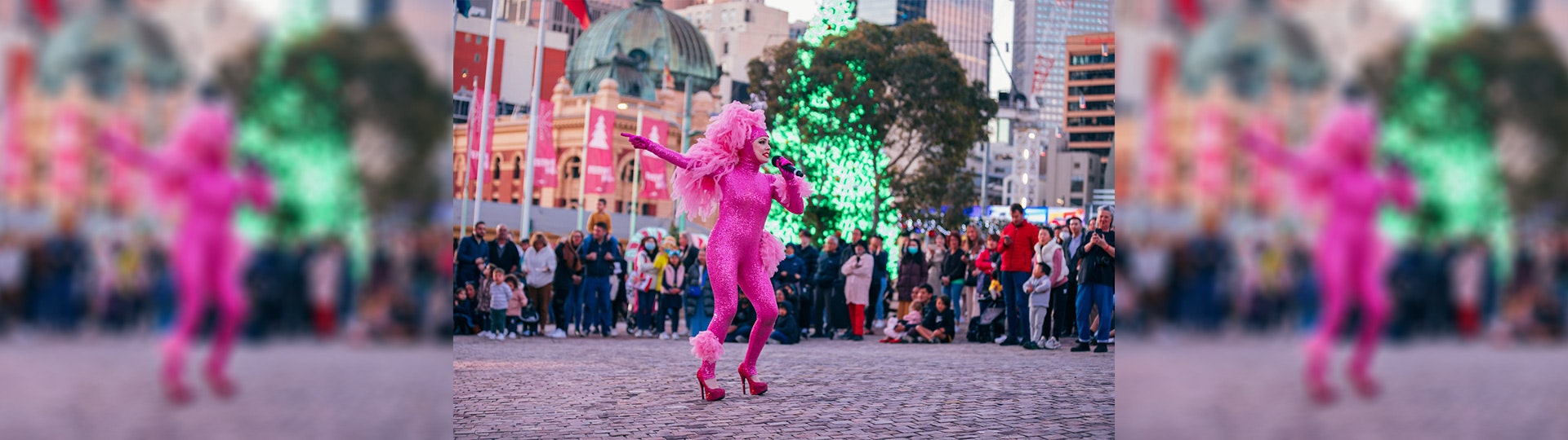 A performer dressed head to toe in bright pink sequins with Flinders St Station and a bright green lit up Christmas tree, on the ground at Fed Square singing