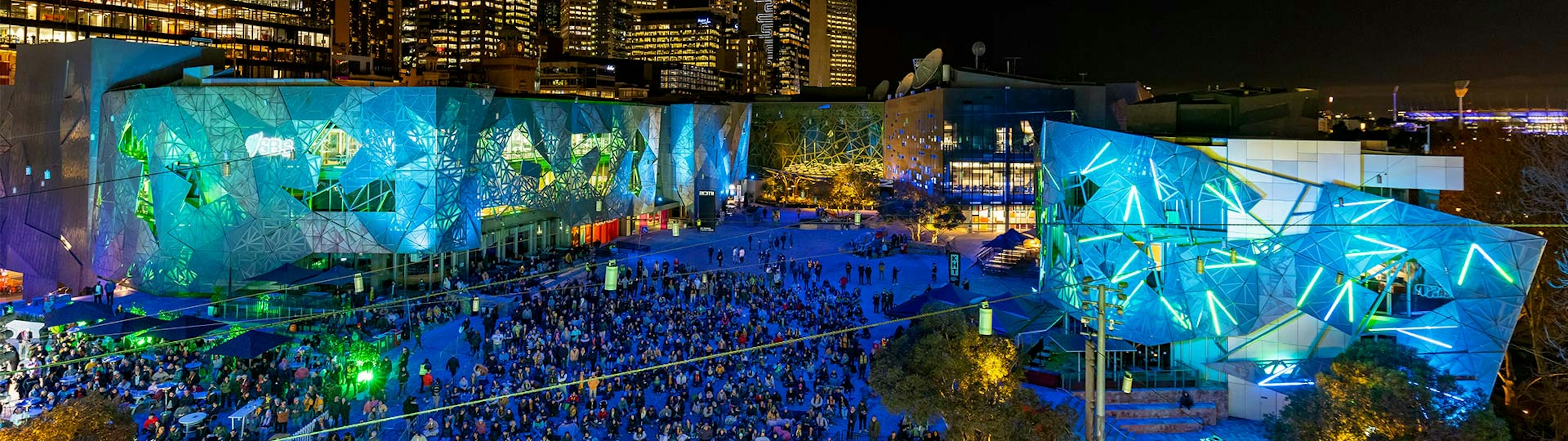 A photo from the top of Fed Square looking down into the Main Square, the square is lit up in blue and green