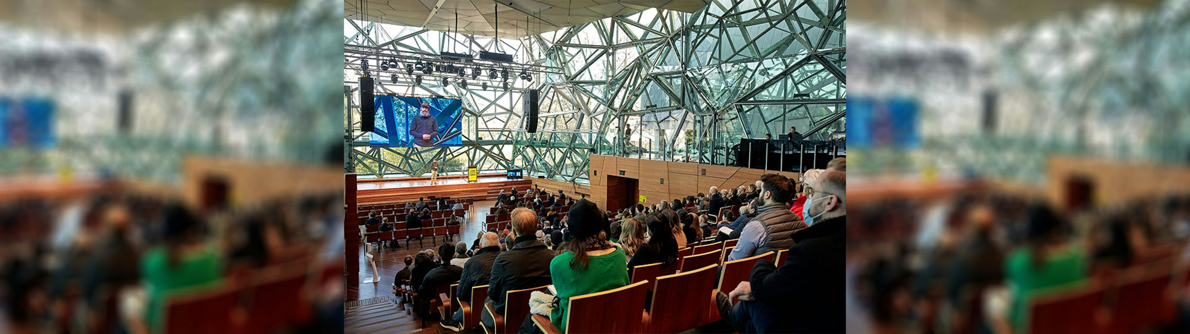 A crowd of people watching a speaker on stage in The Edge, the photo is taken from behind the crowd on the top left hand side