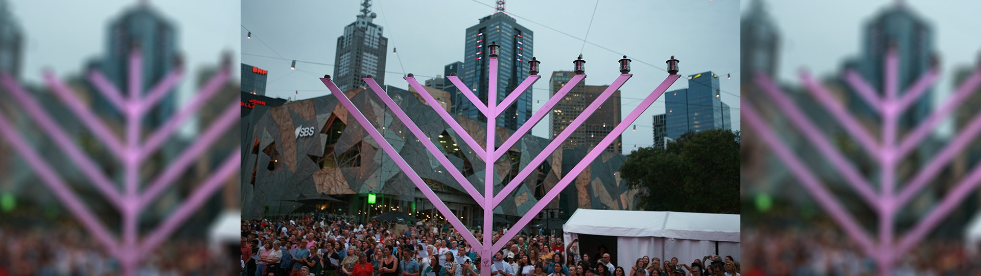 A large metal menorah on the stage at Fed Square with a crowd in the background