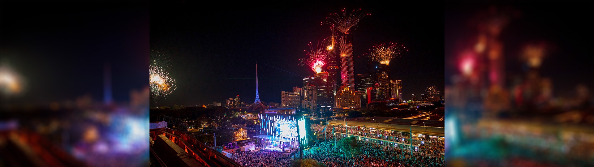 A photo from the top of a building looking down into Fed Square with the crowd watching the big screen. Fireworks can be seen coming off buildings to the south in the distance