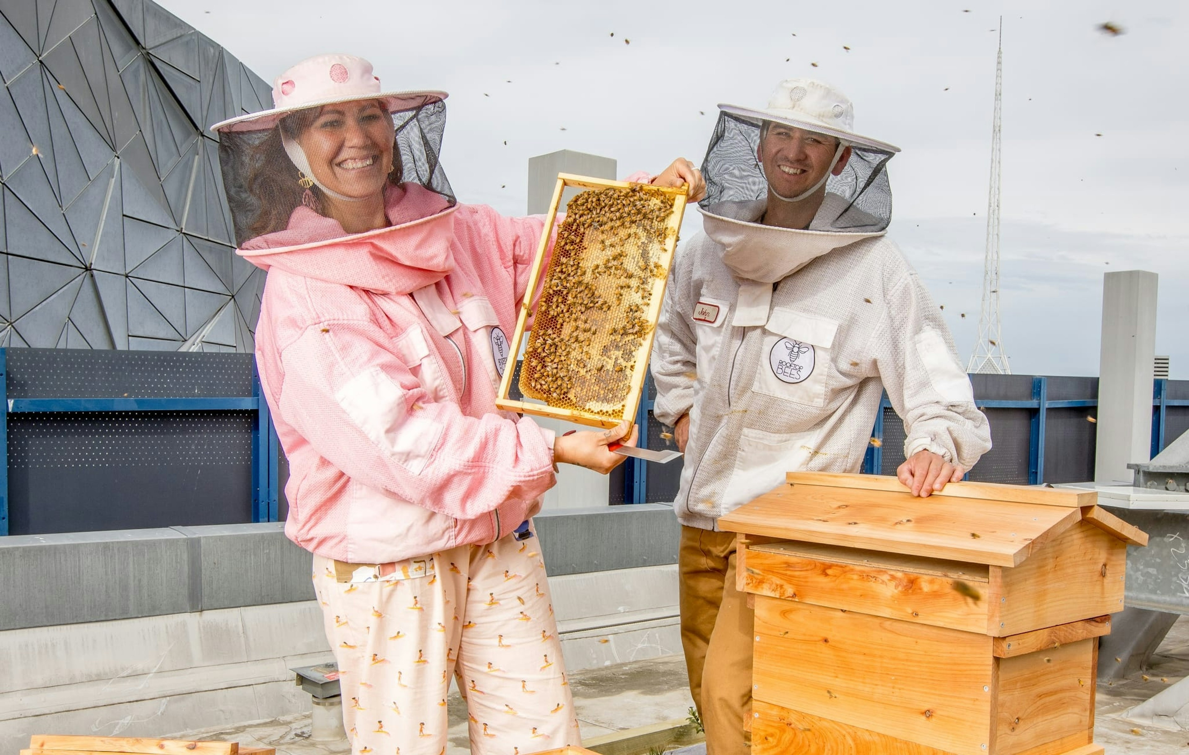 A man and a woman in beekeepers suits stand over two hives on the roof of Fed Square. The woman holds a beehive frame towards the camera, full of bees and honey. The Arts Centre spire is in the background.