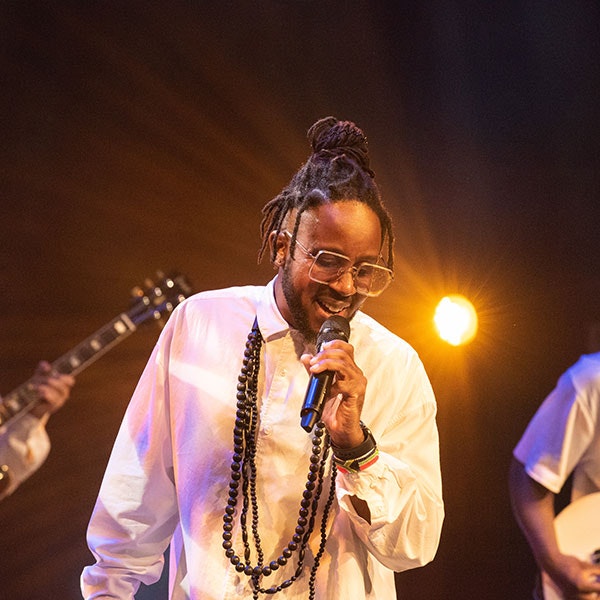 Brotha Asanti is wearing a white shirt with long beads around his neck while singing into a microphone