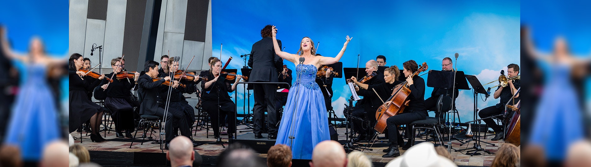 An opera singing is standing on stage with their arms outstrechted in a blue gown, approximately 16 orchstral musicians can be seen behind the singer with a conductor directly behind the singer with their back to the crowd