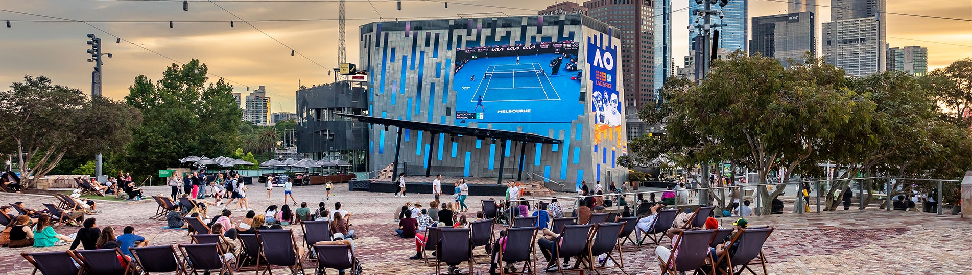 A photo from behind of a crowd of people sitting on deck chairs in Fed Square watching the Australian Open on the Big Screen