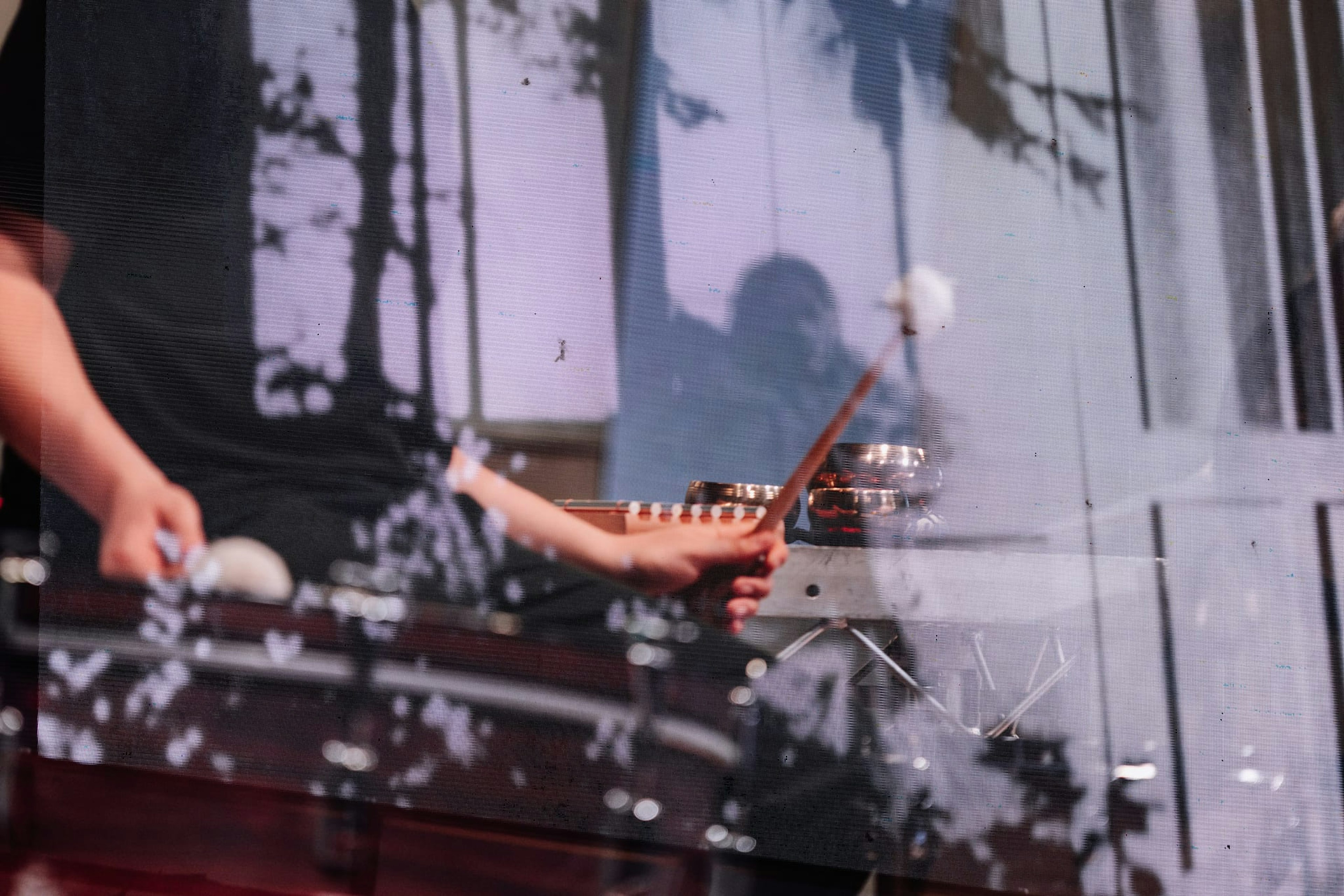 A black and white film is in the background, with a silhouetted figure and palm trees. In front of the screen, hands are raised over a drum, with muted sticks.