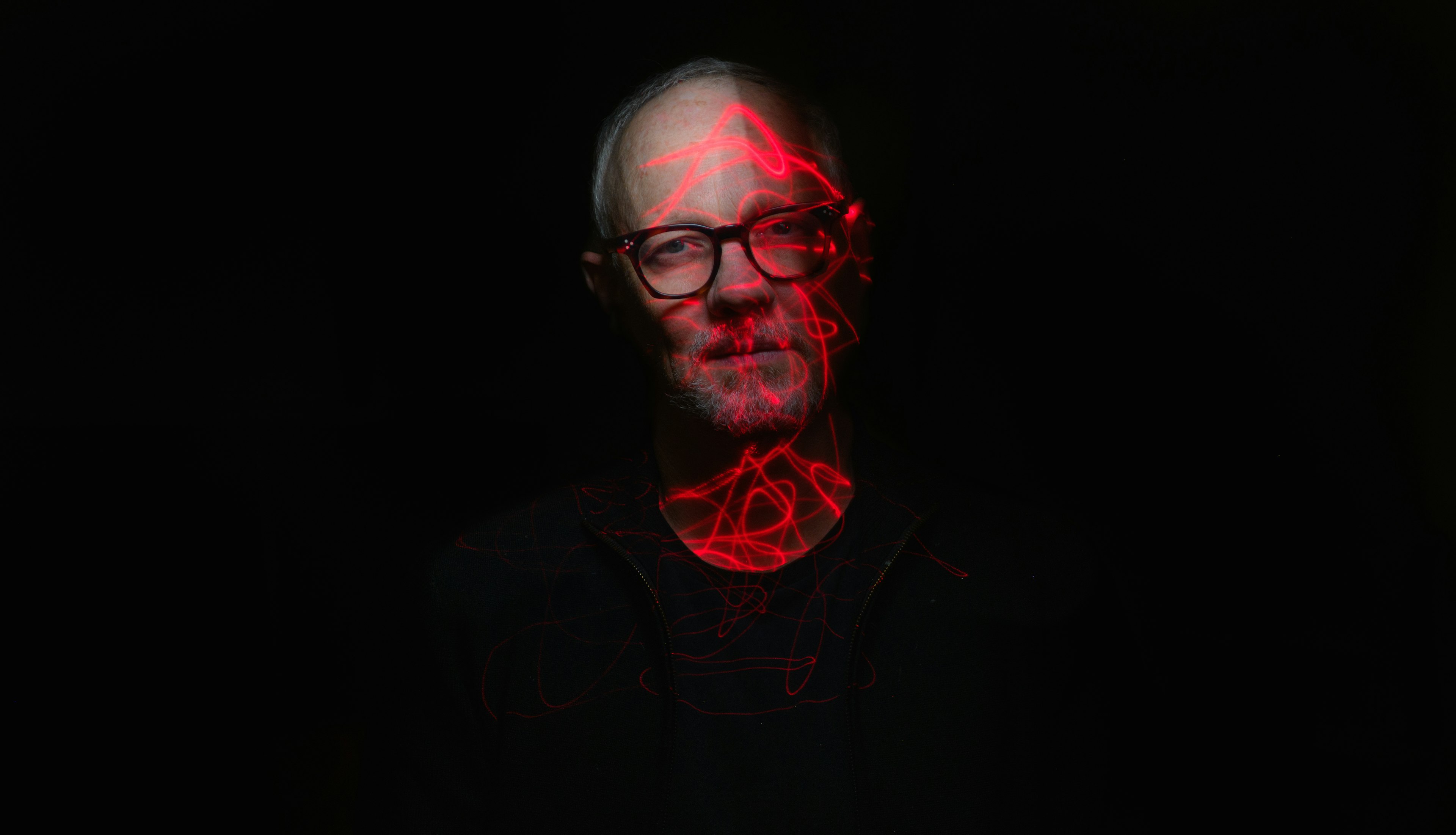 A man with dark-rimming glasses, wearing a black top, looks to the camera, emerging from darkness. Red light forms a pattern across his face.